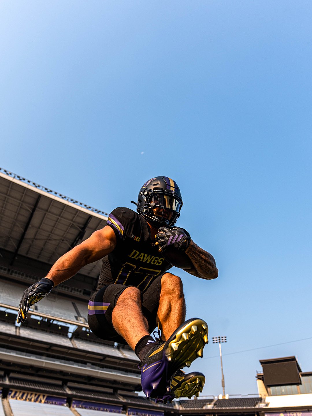 Washington player in the 2026 all-black uniform jumping at Husky Stadium, showing the black pants with purple and gold striping and gold cleats