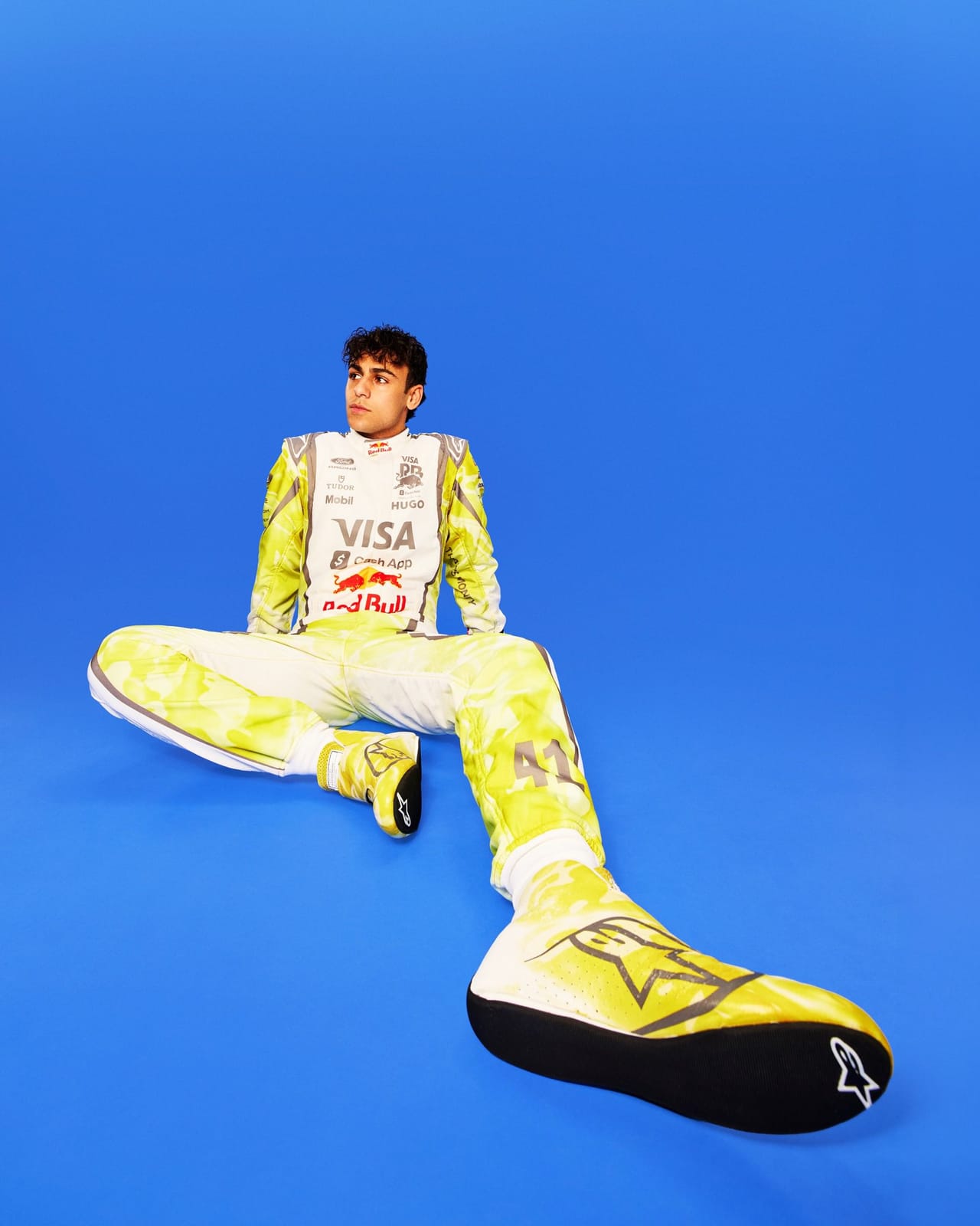 Visa Cash App RB Summer Edition Miami GP 2026 rookie driver Arvid Lindblad in yellow camouflage race suit with white shoulders Red Bull and Visa Cash App branding seated on a deep blue studio background with the number 41 visible on the suit leg