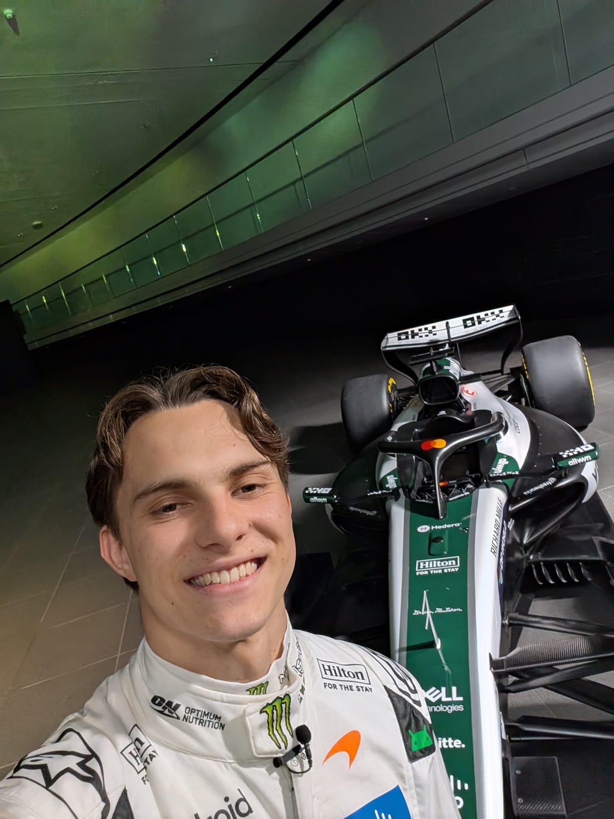McLaren driver Oscar Piastri taking a selfie next to the MCL40 in its 2026 British Grand Prix white-and-green heritage livery, wearing the matching white race suit with the Spark What's Next campaign branding