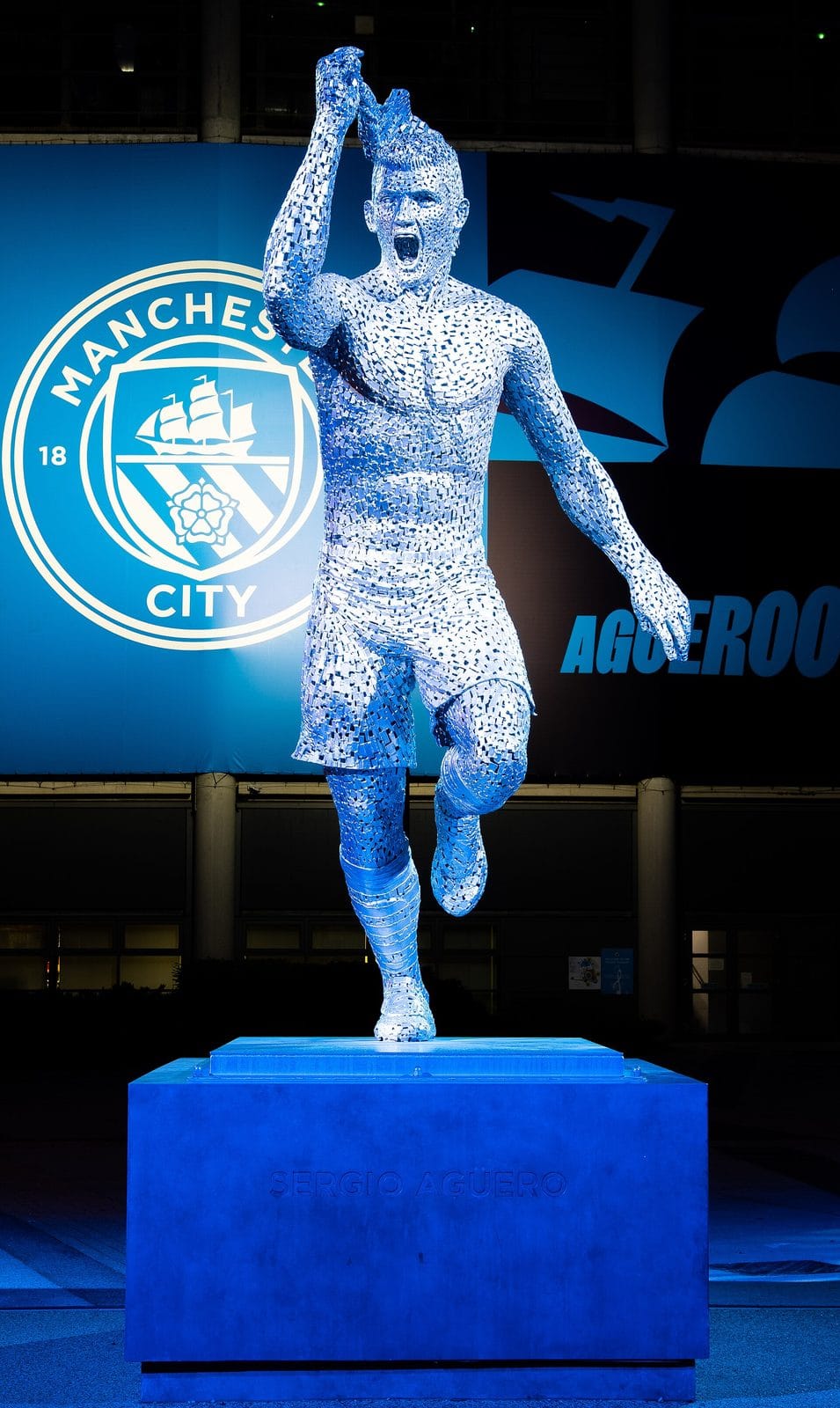 The Manchester City crest, with its ship and three diagonal river stripes, lit up behind the Sergio Aguero statue outside the Etihad Stadium