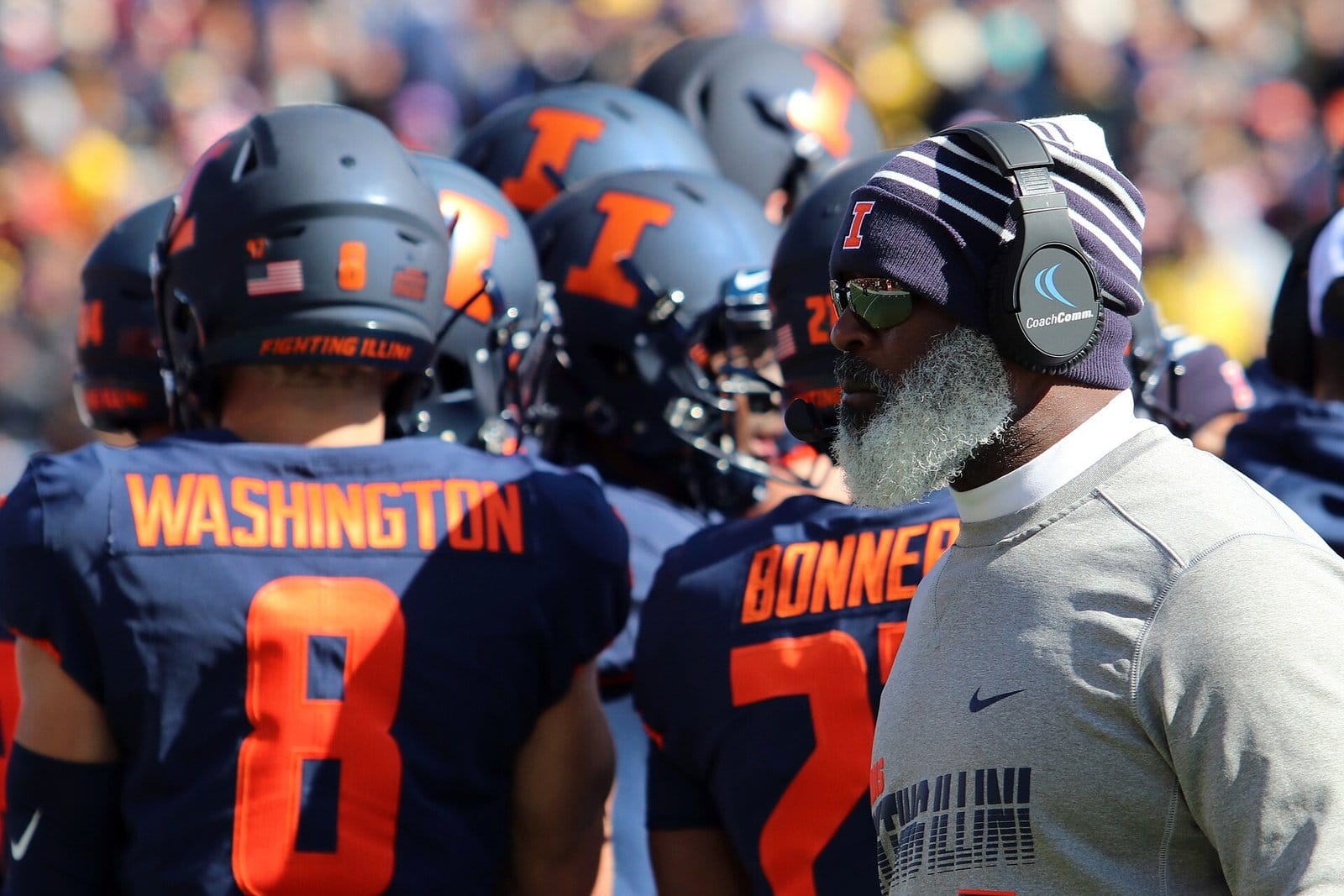 Illinois players in navy home jerseys with orange names and numbers and gray helmets on the sideline at Memorial Stadium