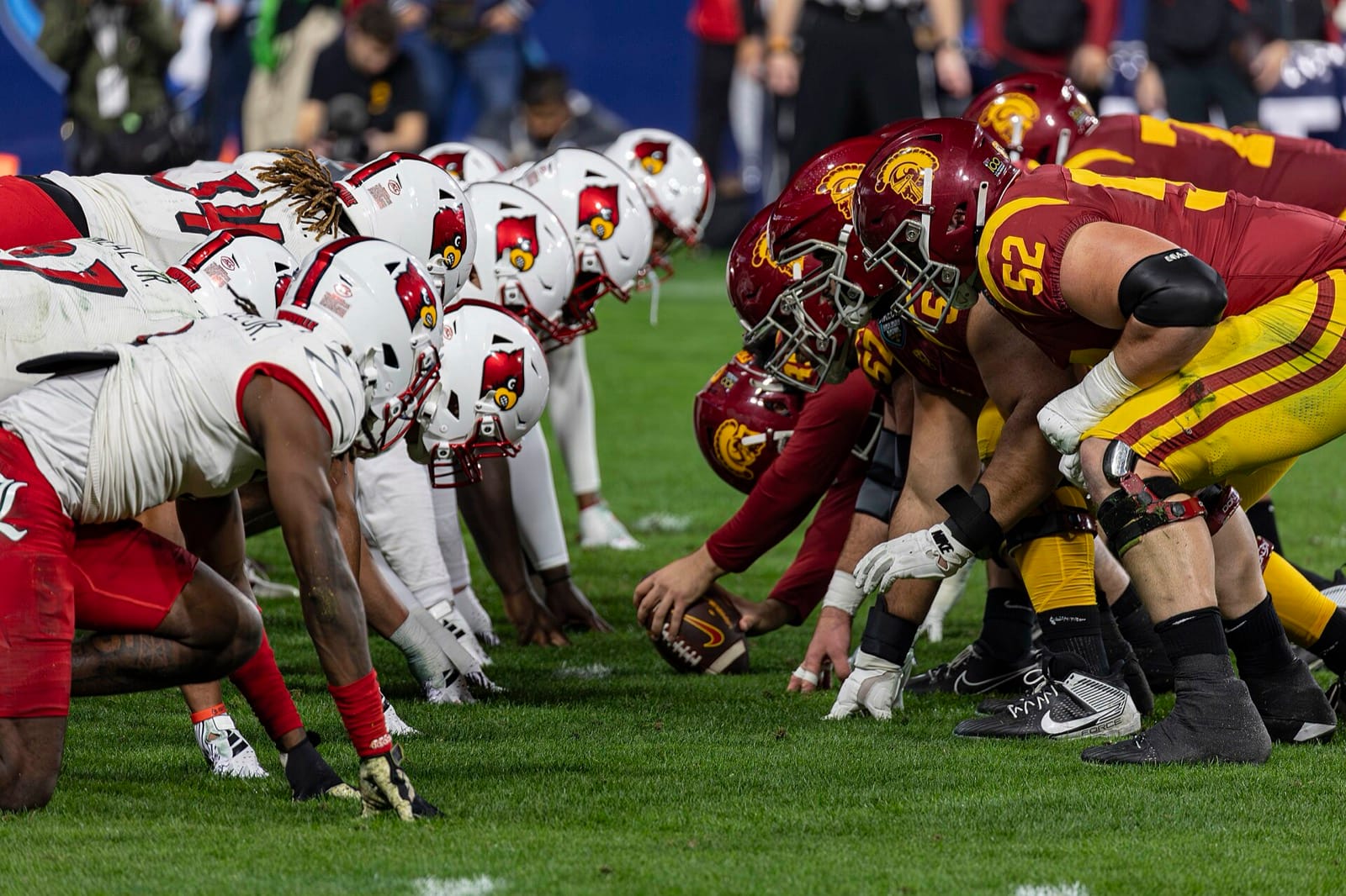 Louisville linemen in white jerseys with red trim and cardinal-logo helmets at the line against USC at the 2023 Holiday Bowl