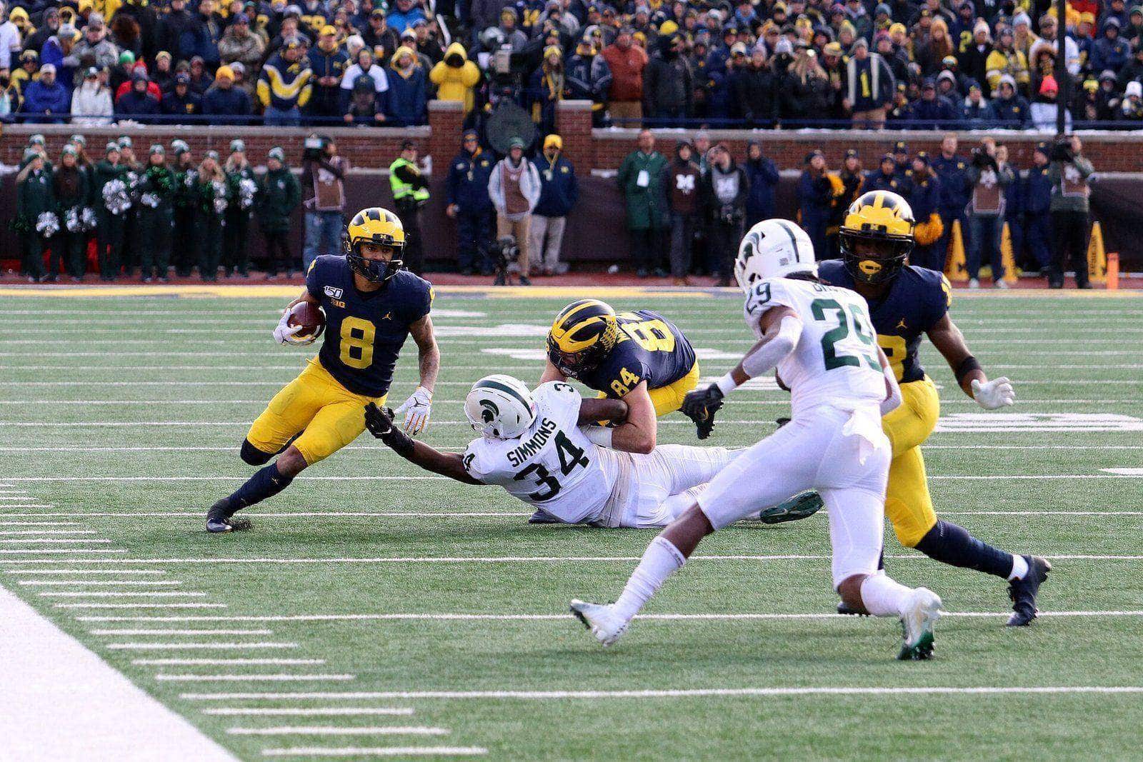 A Michigan player in the navy home jersey, maize pants and winged helmet runs with the ball as Michigan State defenders chase