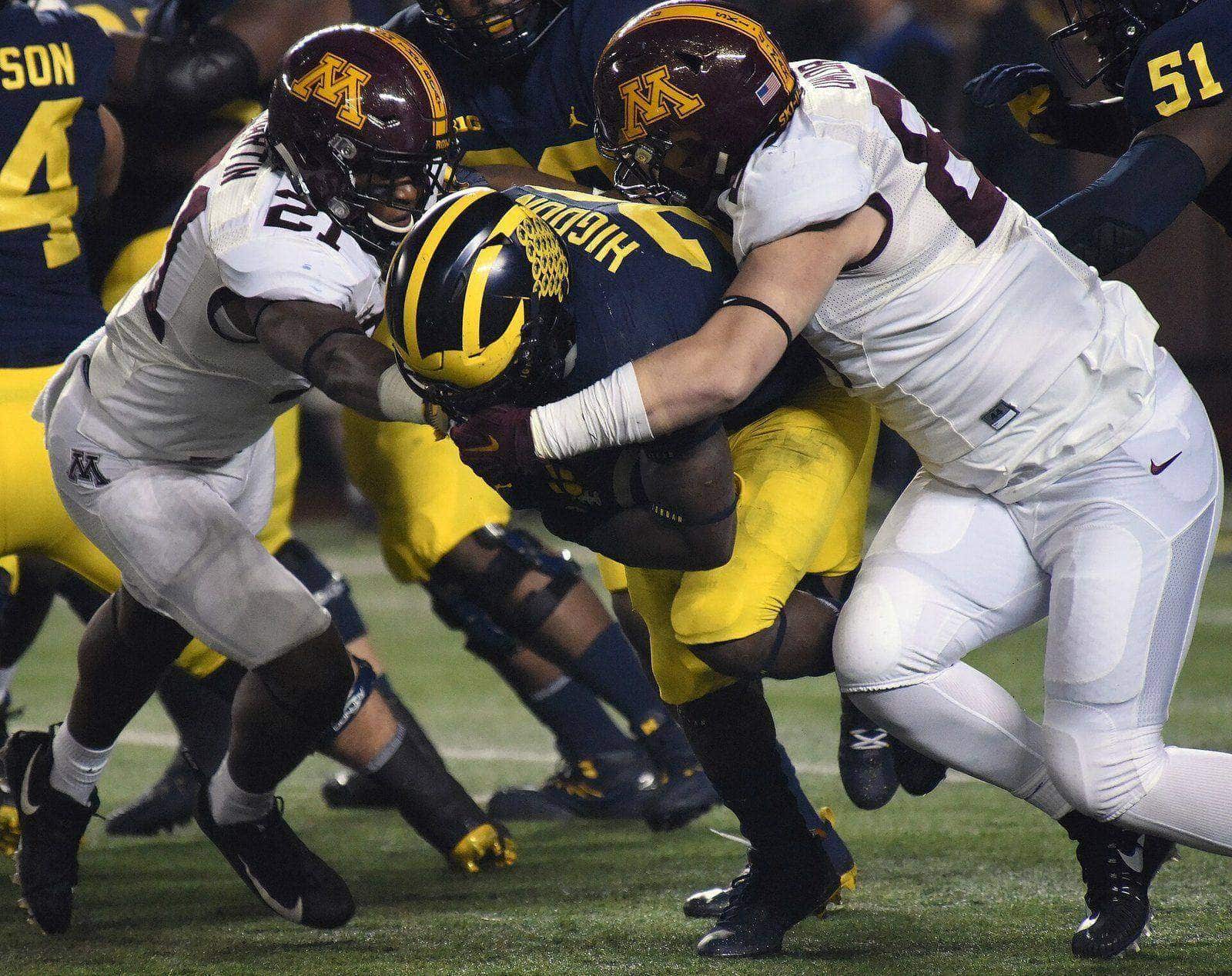 Minnesota defenders in white road jerseys and maroon helmets with the gold M make a tackle at Michigan Stadium