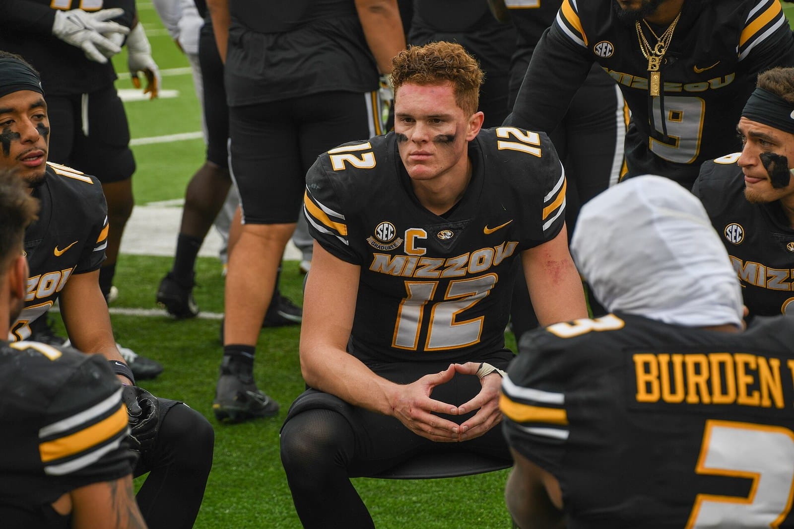 Missouri quarterback Brady Cook and teammates in black jerseys kneel on the sideline at Faurot Field