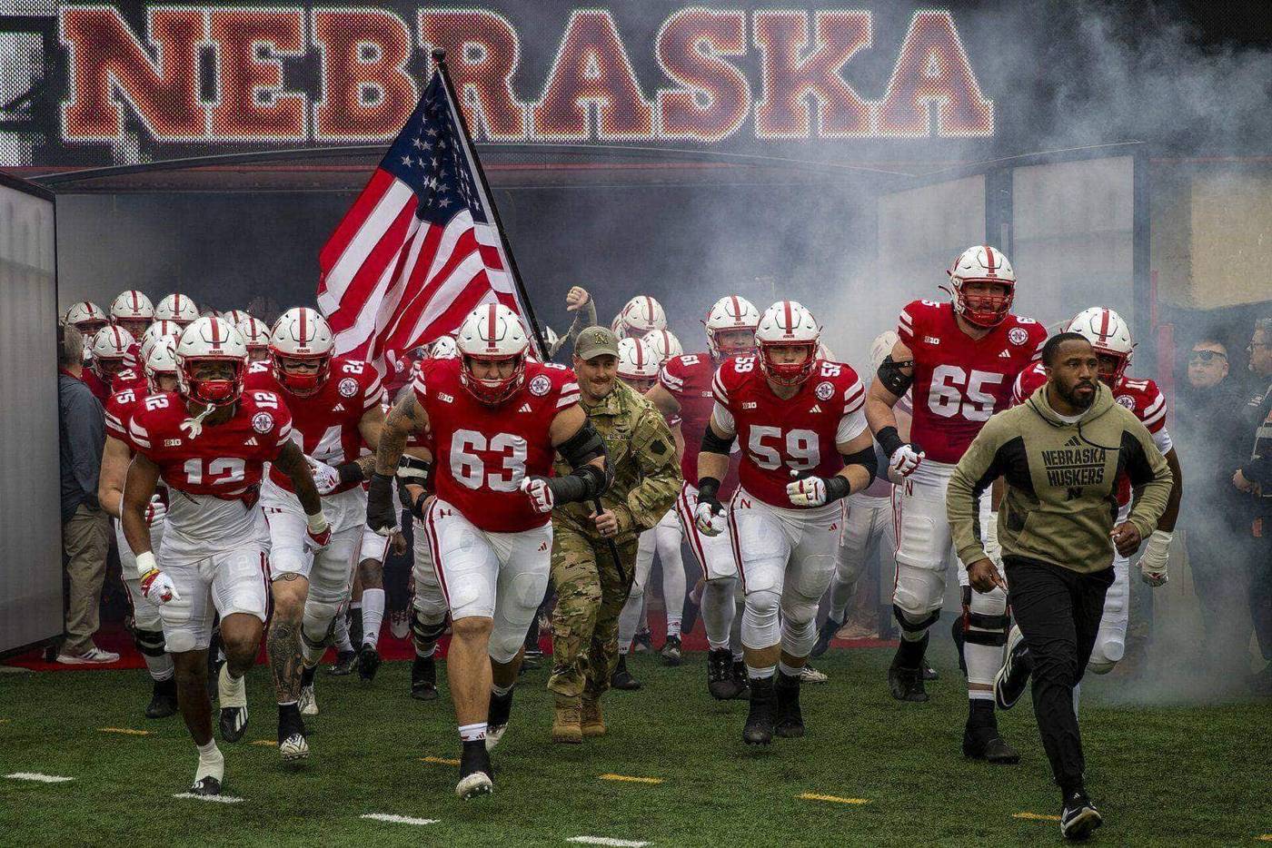 Nebraska players in red home jerseys and white helmets run out of the tunnel under the NEBRASKA sign at Memorial Stadium