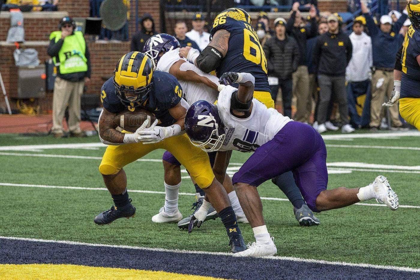 Northwestern defenders in white road jerseys, purple pants and purple helmets tackle a Michigan ball carrier at the goal line