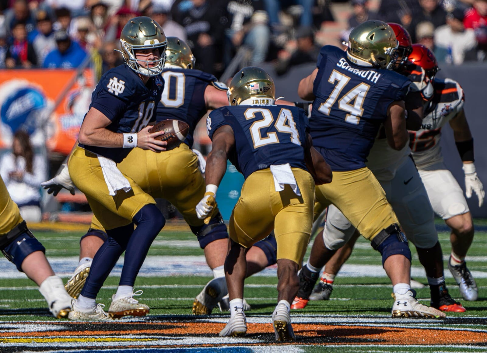 Notre Dame in navy jerseys, gold pants and gold helmets as a ball carrier runs behind blockers at the 2023 Sun Bowl