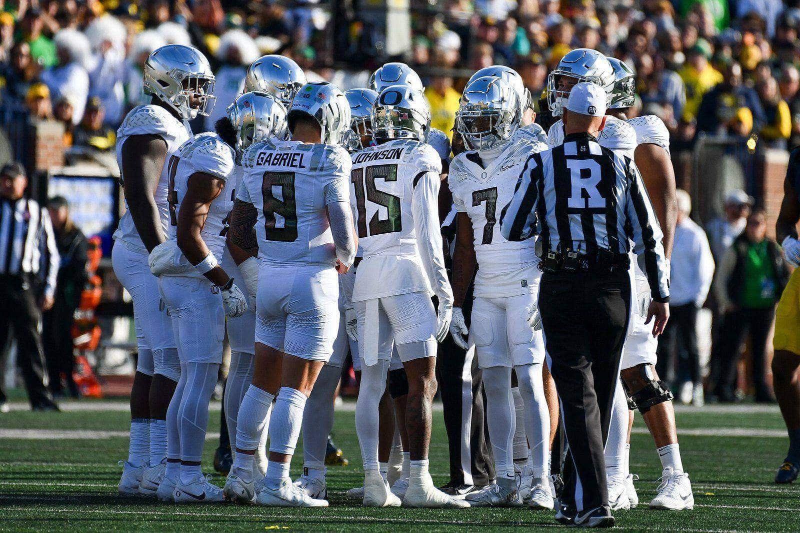 Oregon's offense in a huddle in all-white road uniforms with chrome helmets at Michigan Stadium