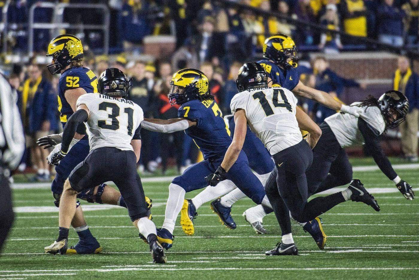 Purdue players in white road jerseys, black pants and black helmets chase a Michigan ball carrier at night