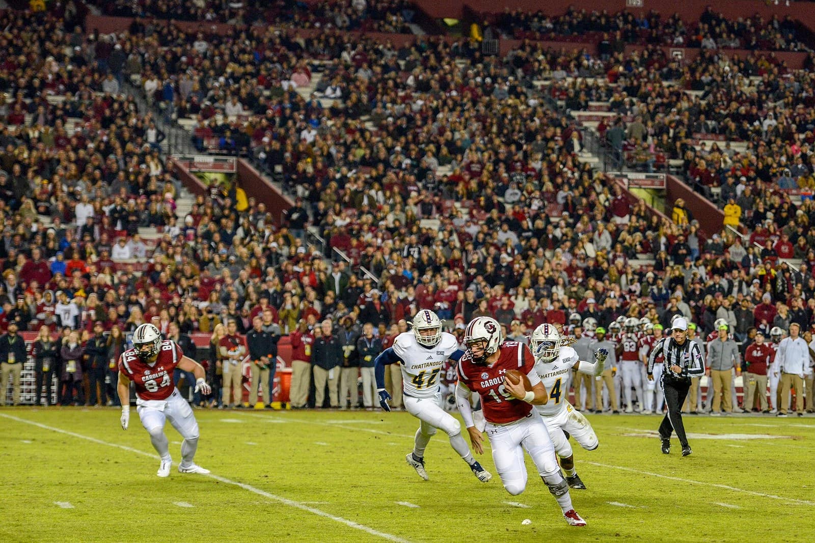South Carolina players in garnet jerseys and white helmets run a play at night at Williams-Brice Stadium