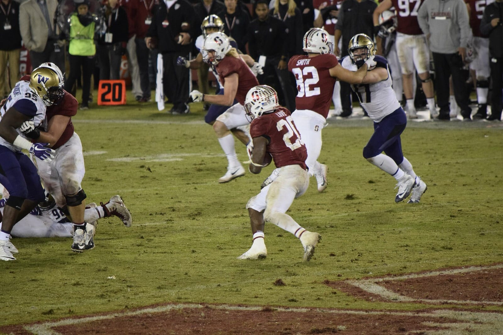 A Stanford ball carrier in a cardinal jersey and white helmet runs past Washington defenders at Stanford Stadium