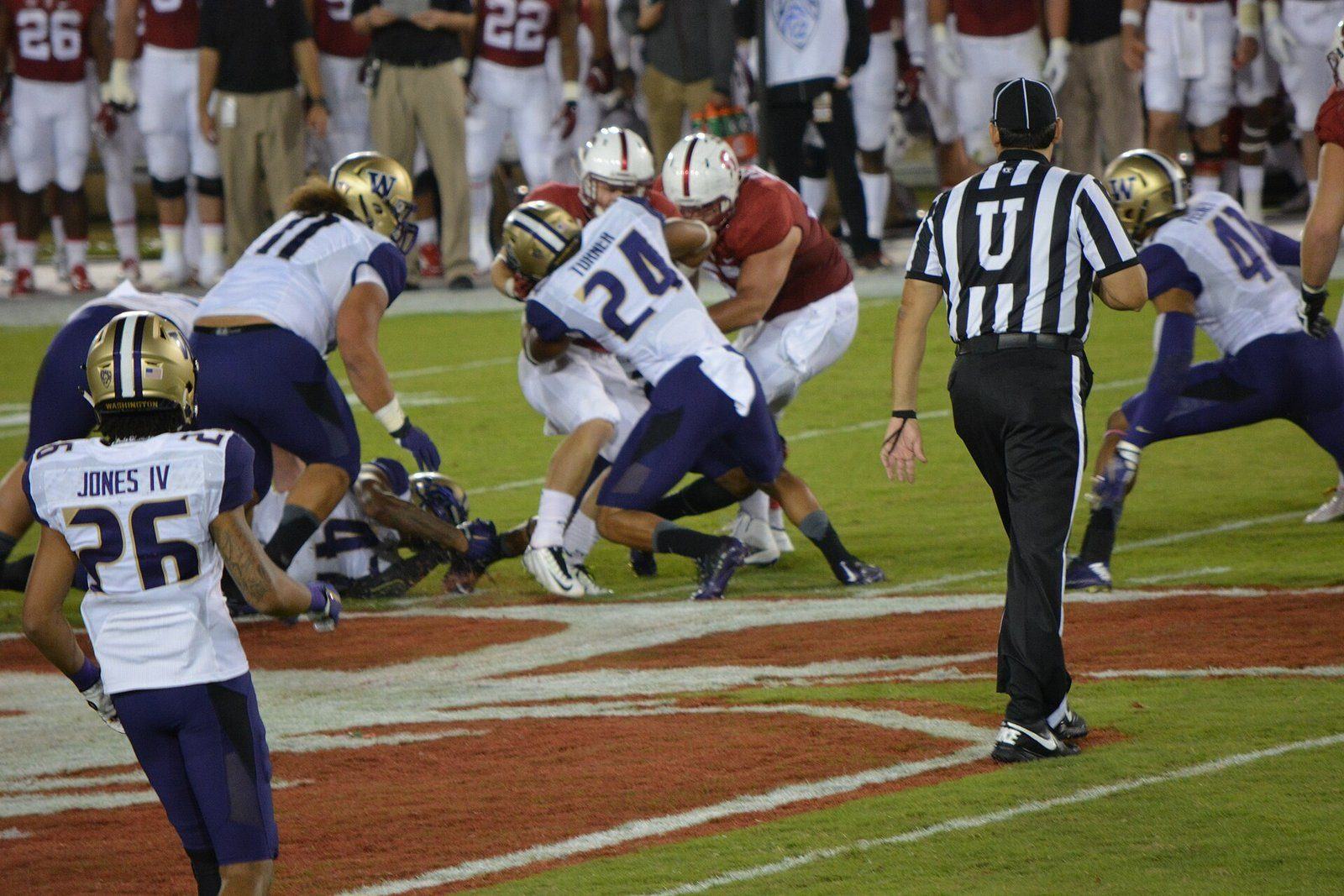 Washington defenders in white road jerseys, purple pants and gold helmets tackle a Stanford ball carrier