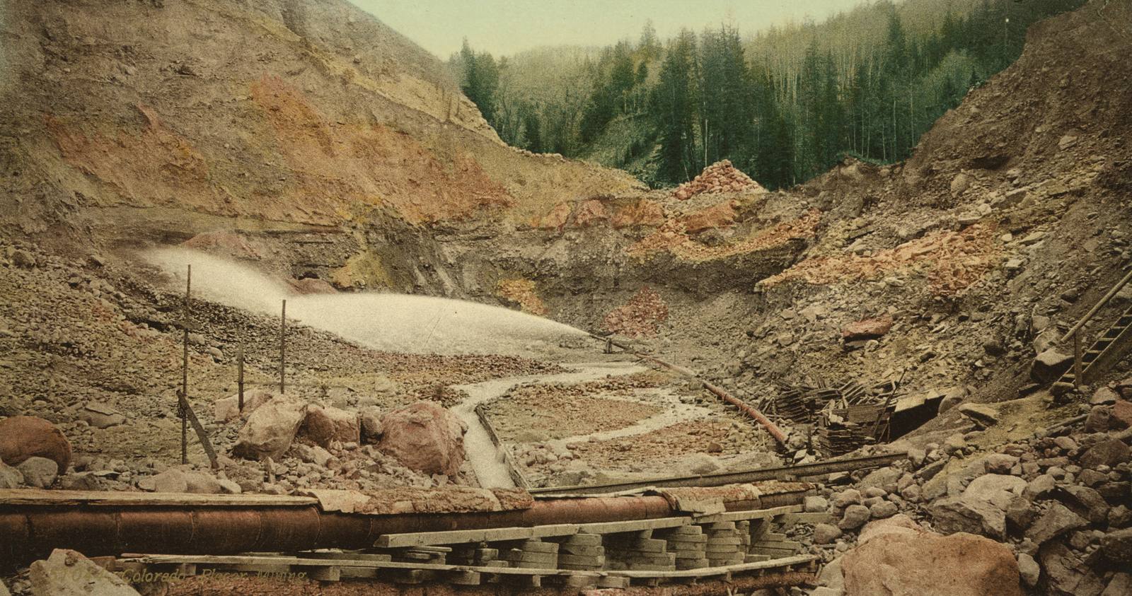 A Colorado placer mining site around 1900, a water jet washing a gravel bank with pipes and sluices in the foreground