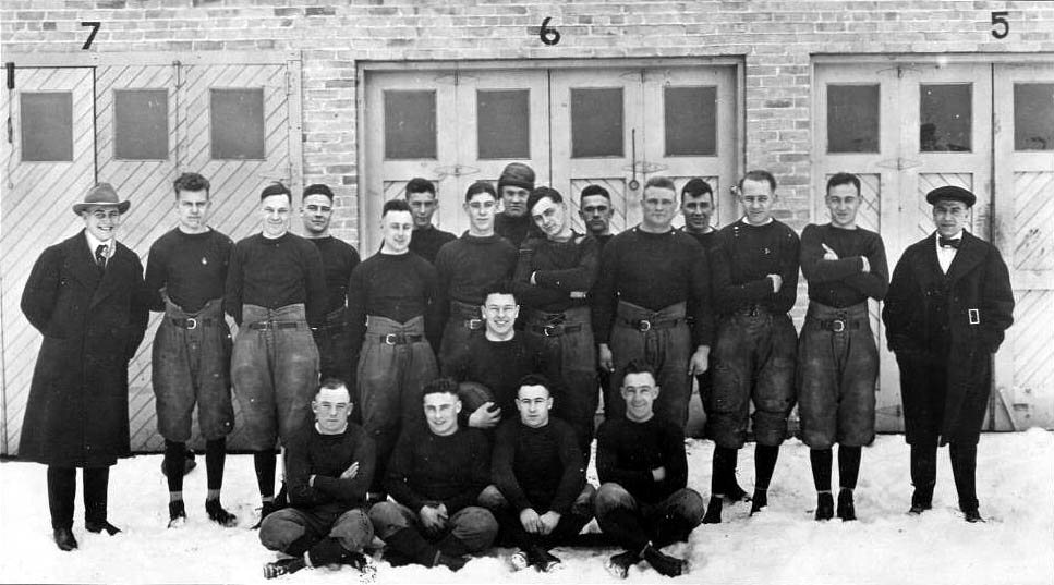 The 1919 Green Bay Packers pose in plain dark jerseys outside the Indian Packing Company building, with Curly Lambeau among them (black-and-white photo)