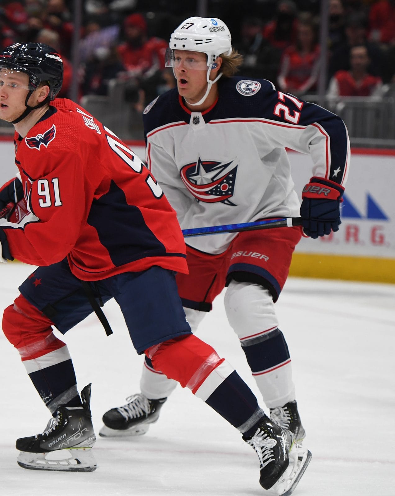 Adam Boqvist in the Blue Jackets' white road uniform with the star-and-flag crest and navy and red trim, No. 27, skates beside the Capitals' Joe Snively in red at Capital One Arena in Washington, February 8, 2022