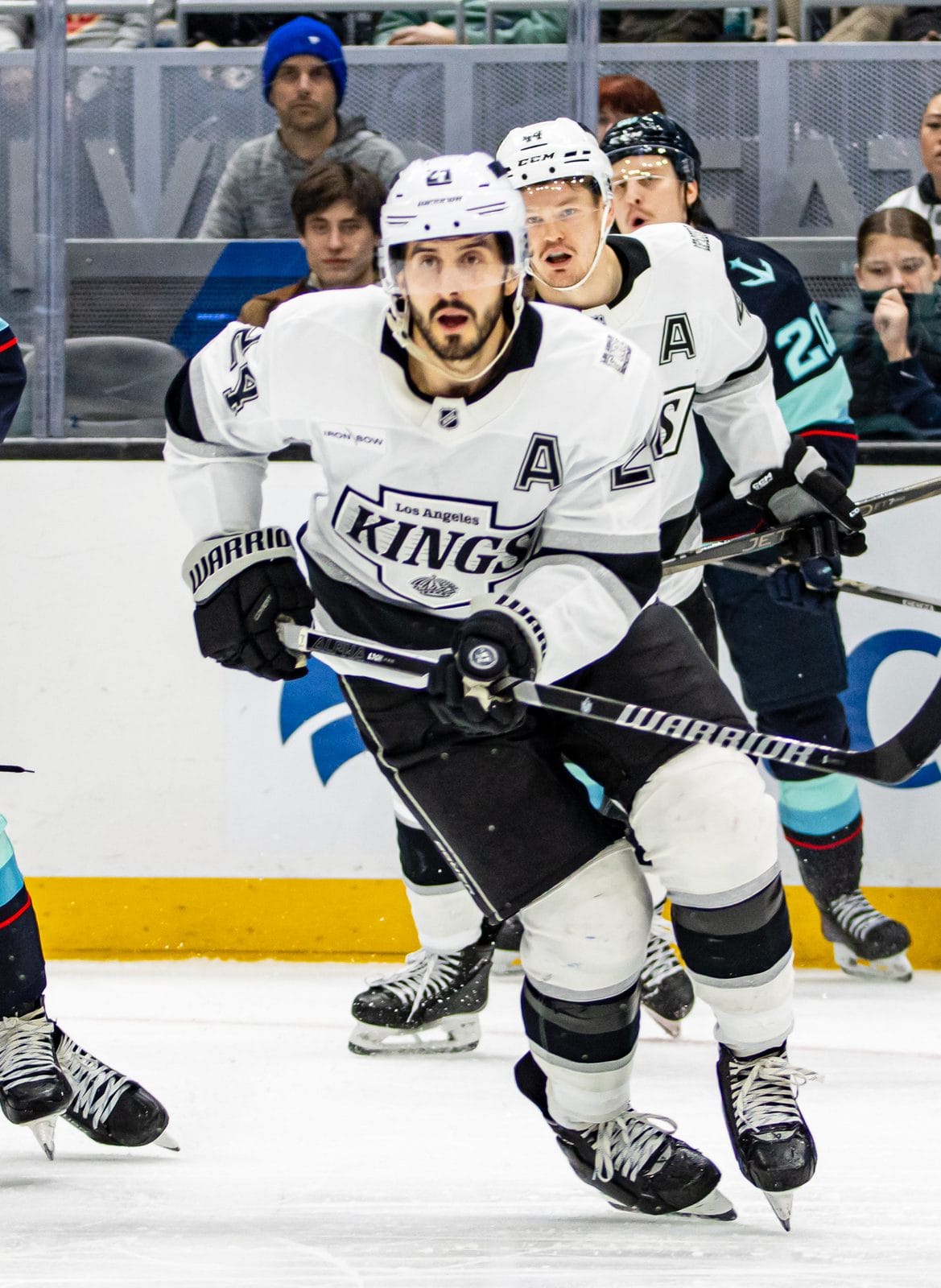 Phillip Danault, wearing an A, in the Kings' white road uniform with the black-and-silver KINGS crest skates up ice beside the Kraken's Shane Wright at Climate Pledge Arena in Seattle, January 18, 2025