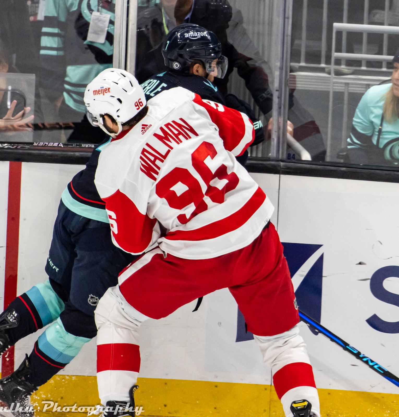 Jake Walman in the Red Wings' white road uniform with red shoulders and WALMAN over No. 96 pins a Kraken player to the boards at Climate Pledge Arena in Seattle, February 18, 2023