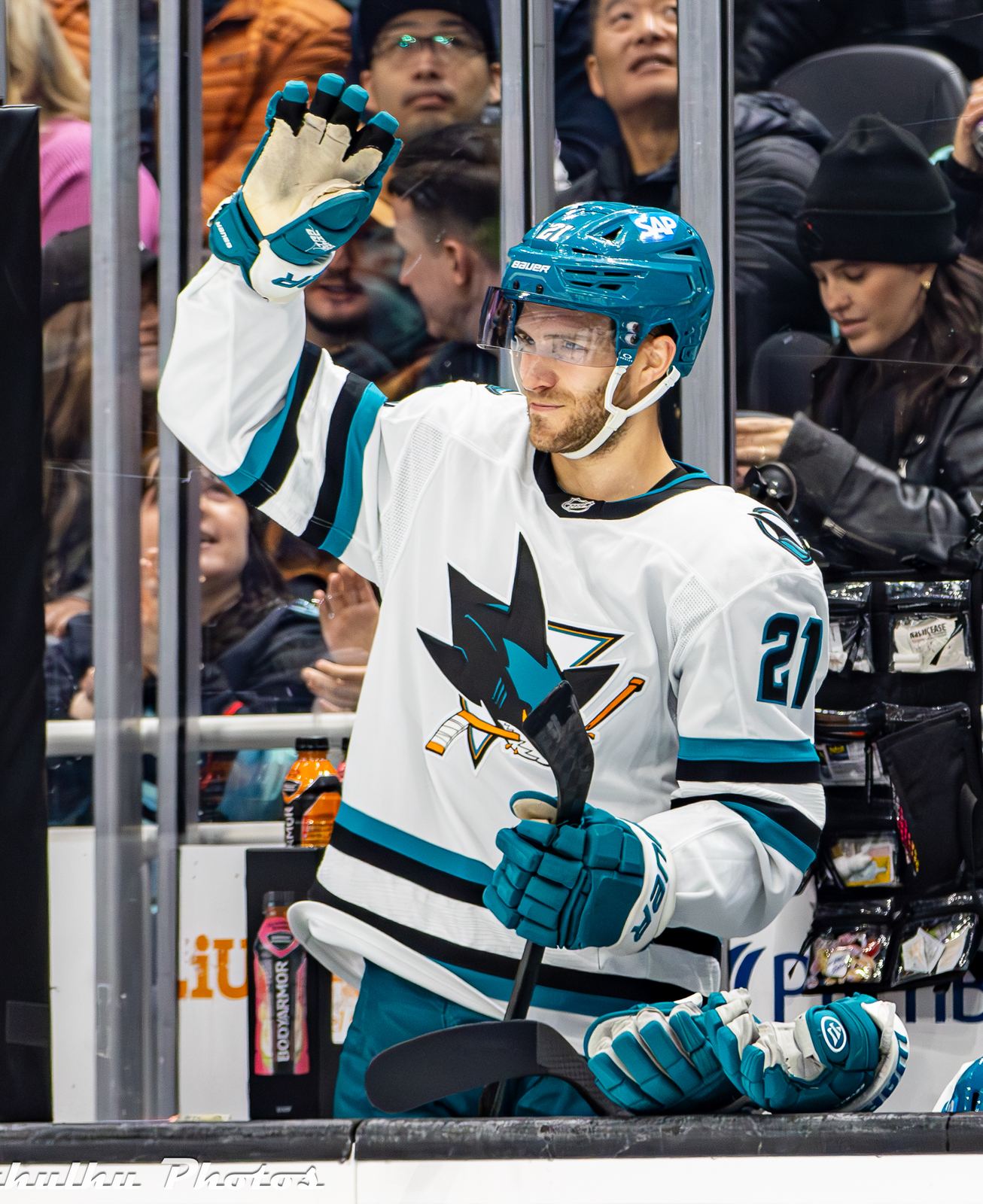 A Sharks player in the white road uniform with teal trim and the shark-and-stick crest, No. 21, waves from the bench at Climate Pledge Arena in Seattle, November 30, 2024