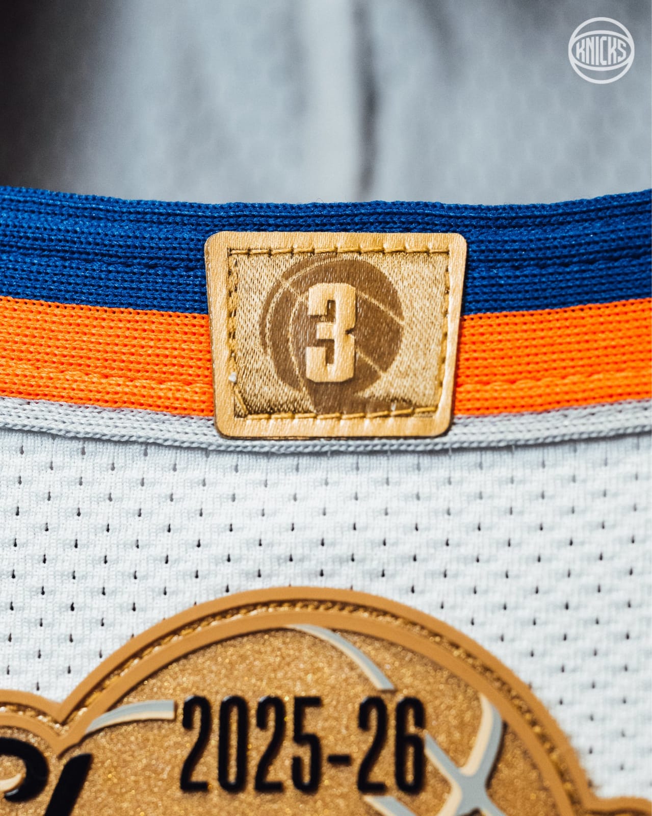 Close-up of the gold 3 tag at the back collar of the Knicks ring night jersey, with the blue and orange collar stripes and the top of the gold 2025-26 Champions patch below