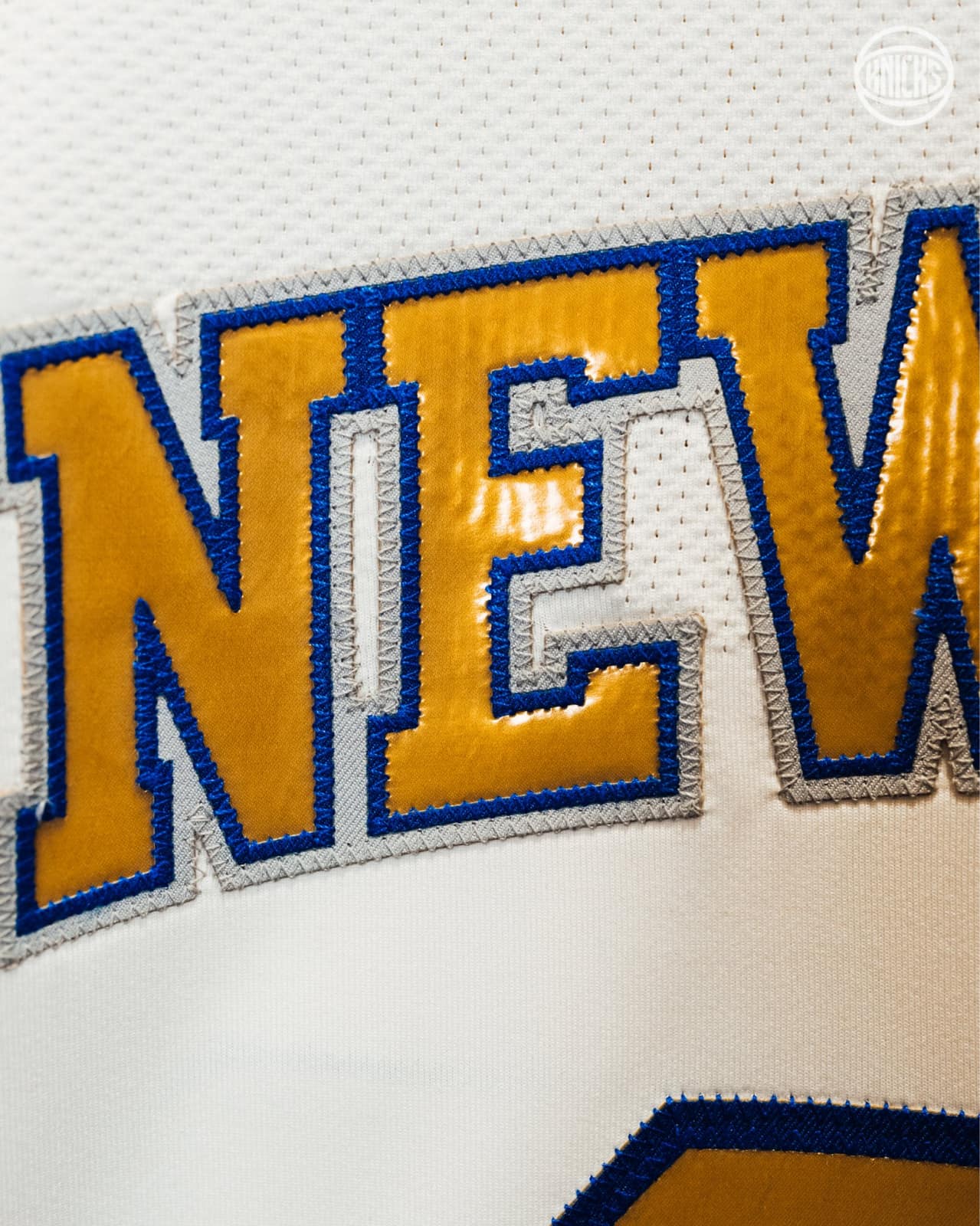 Close-up of the Knicks ring night jersey front: NEW YORK wordmark in metallic gold with blue and gray outlines on white mesh