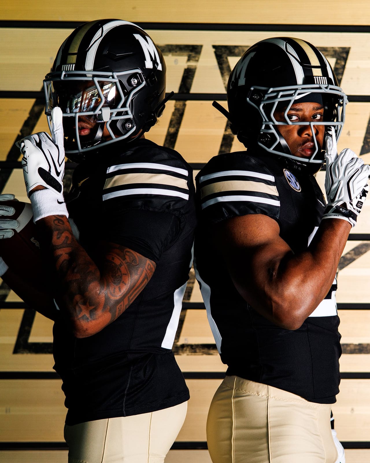 Two Missouri players in the 2026 Memorial Stadium 100th anniversary throwback uniform, black helmets with a gold and white center stripe and block M, black jerseys with gold and white shoulder stripes, and gold pants