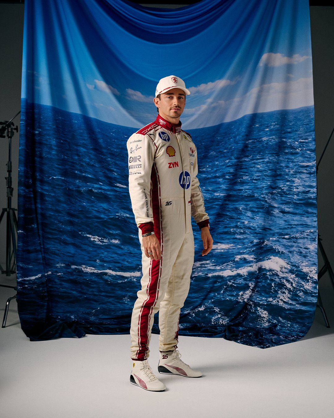 Charles Leclerc Ferrari F1 driver standing in his special Monaco Grand Prix 2026 home race suit, a white base with red vertical stripes inspired by the Monaco flag with the HP title sponsor logo on the chest opposite the Ferrari prancing horse crest, his race number 16 on the leg, and a matching white cap with red Ferrari logo, photographed in front of an ocean and sky backdrop that evokes the Mediterranean coastline of his home principality