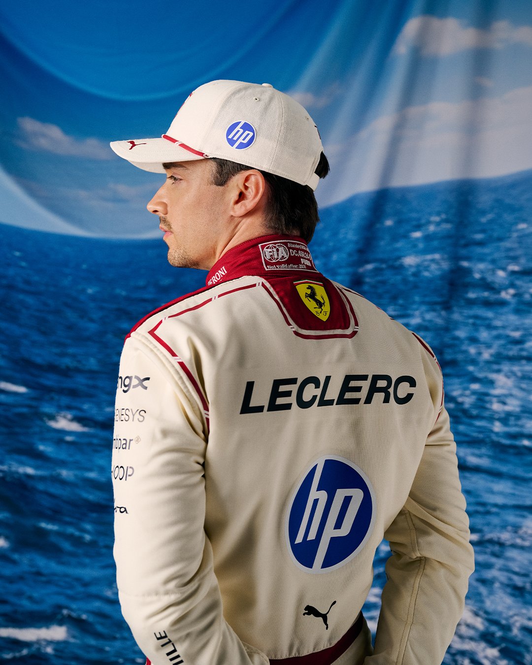 Charles Leclerc Ferrari F1 Monaco Grand Prix 2026 special home race suit pose with the matching white cap featuring the red Ferrari roundel logo and the ocean and Mediterranean coastline backdrop