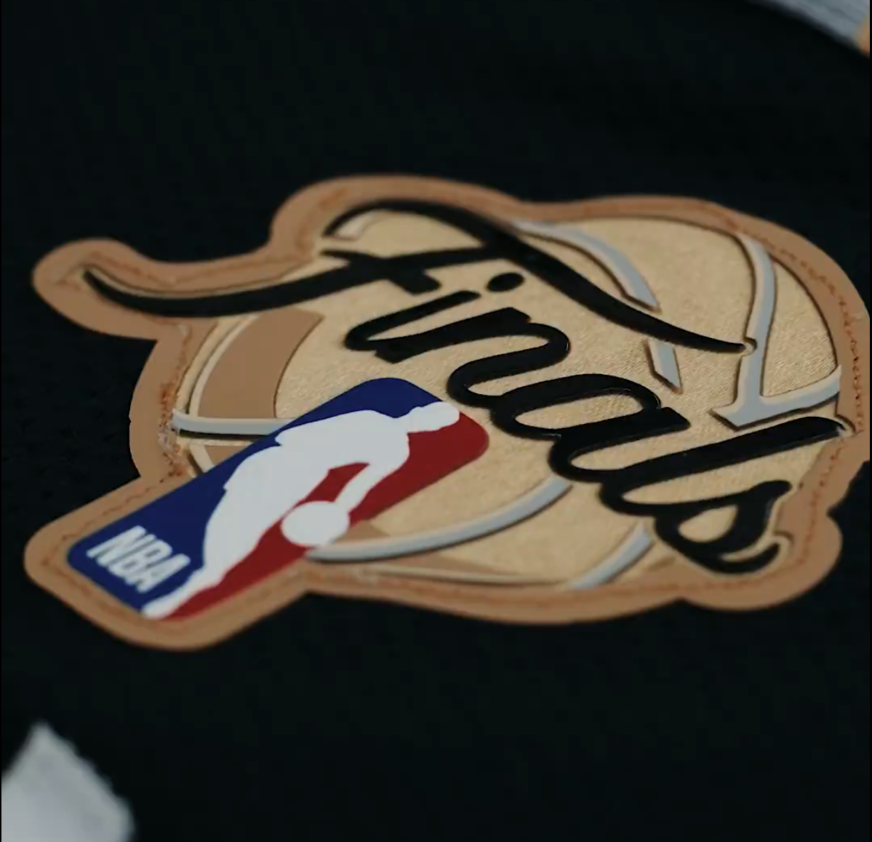 Close-up macro shot of the 2026 NBA Finals back patch hand-stitched onto the Spurs Icon Edition black jersey showing the embroidered tan leather-toned basketball-textured background with the Finals cursive script wordmark across the top, the NBA Jerry West silhouette logo on a red and white and blue tab anchoring the bottom of the patch, and the visible thread stitching detail around the patch border