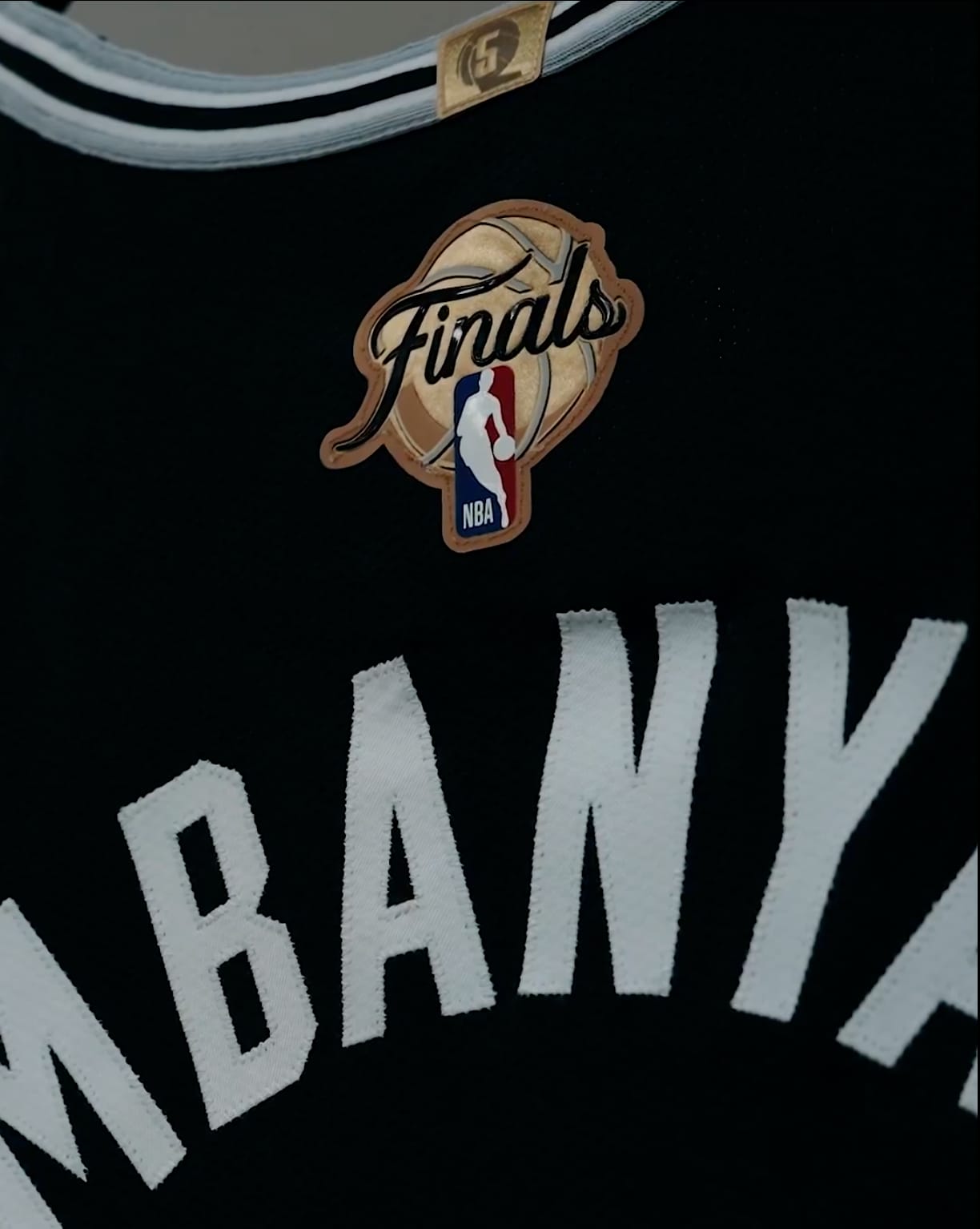 2026 NBA Finals back patch hand-stitched onto the upper back yoke of Victor Wembanyama's San Antonio Spurs Icon Edition black jersey, the patch carrying a tan leather-colored basketball-textured background with the Finals cursive script wordmark across the top, the NBA Jerry West silhouette logo on a red and white and blue tab anchoring the bottom of the patch, and the back nameplate WEMBANYAMA stitched in white block letters directly below the patch