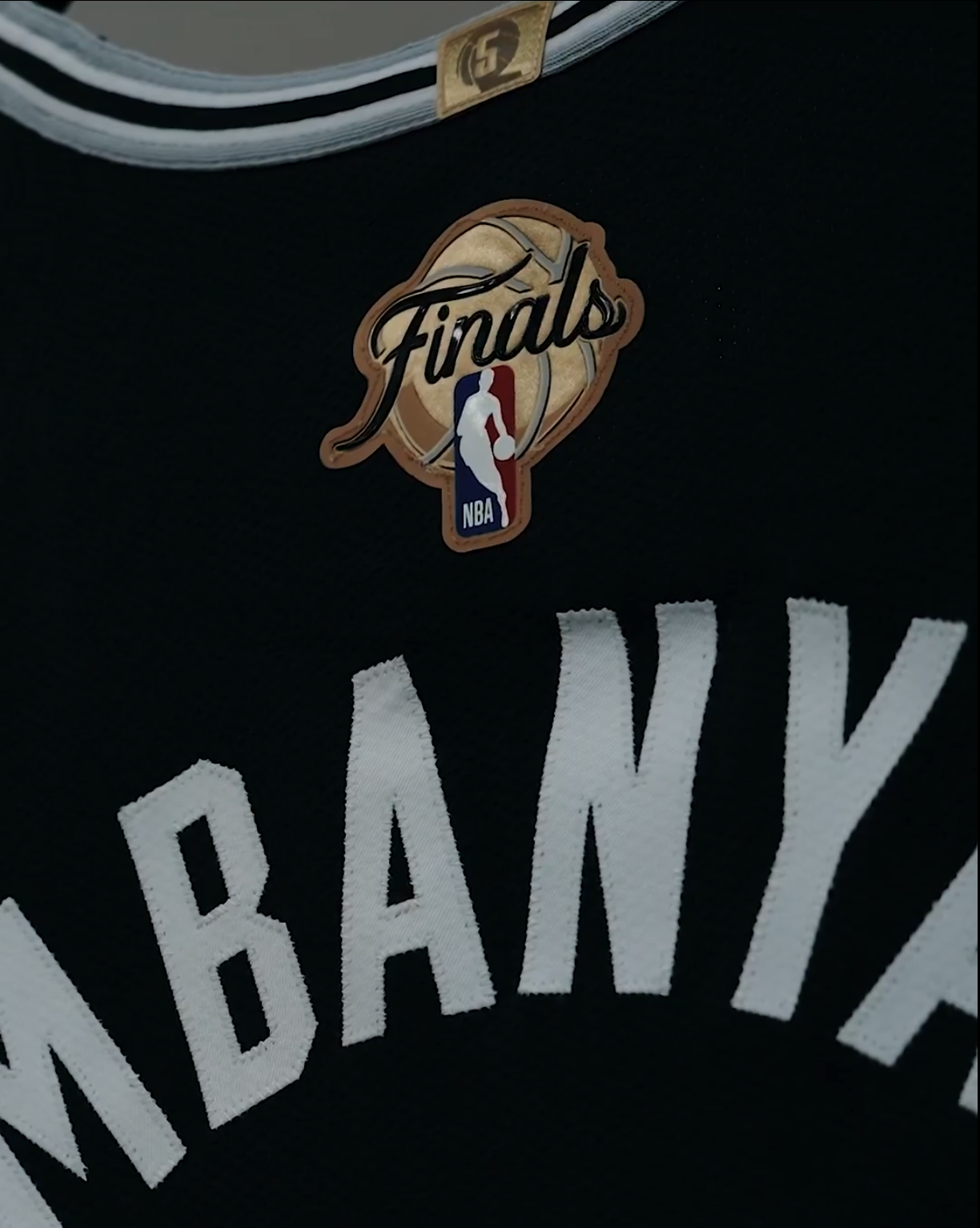 2026 NBA Finals back patch hand-stitched onto the upper back yoke of Victor Wembanyama's San Antonio Spurs Icon Edition black jersey, the patch carrying a tan leather-colored basketball-textured background with the Finals cursive script wordmark across the top, the NBA Jerry West silhouette logo on a red and white and blue tab anchoring the bottom of the patch, and the back nameplate WEMBANYAMA stitched in white block letters directly below the patch