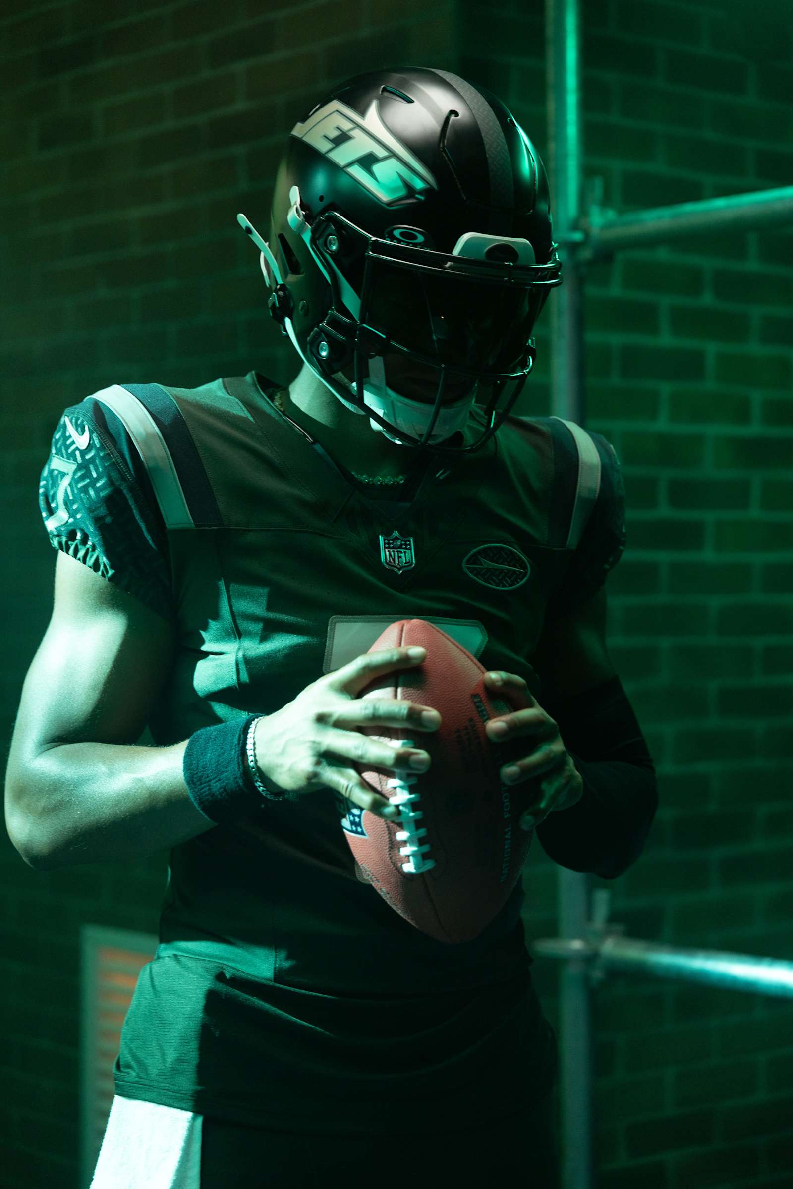 New York Jets Gotham City Football Rivalries uniform, a dark green jersey with UCLA-style shoulder stripes, a black gloss helmet with an italic JETS wordmark, shown on a player holding a football under green light