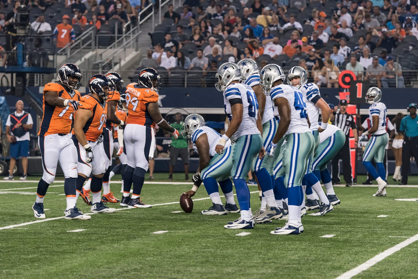 The Dallas Cowboys in white jerseys at the line of scrimmage against the Denver Broncos in orange at AT&T Stadium in Arlington, Texas, the home team in white and the visitors in color