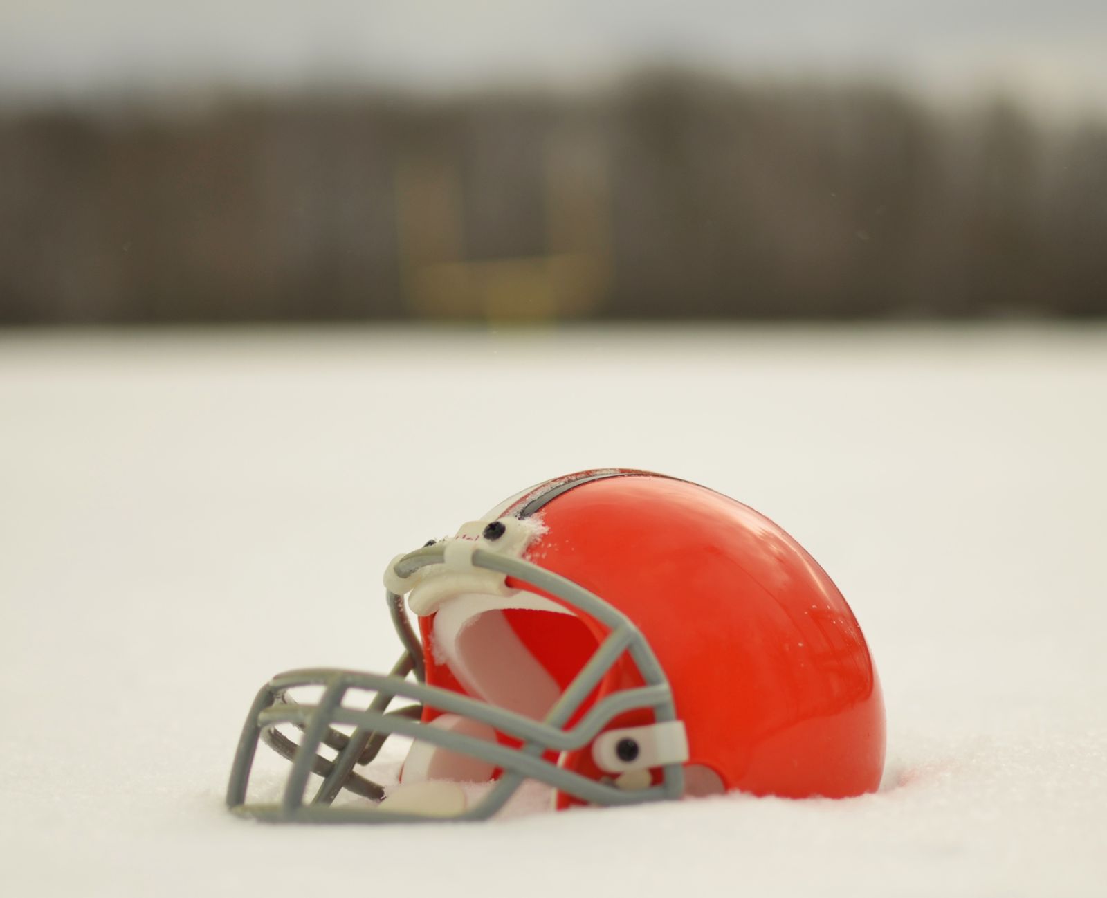 A Cleveland Browns orange helmet with a brown and white center stripe and a gray facemask, sitting in snow, with no logo on the side