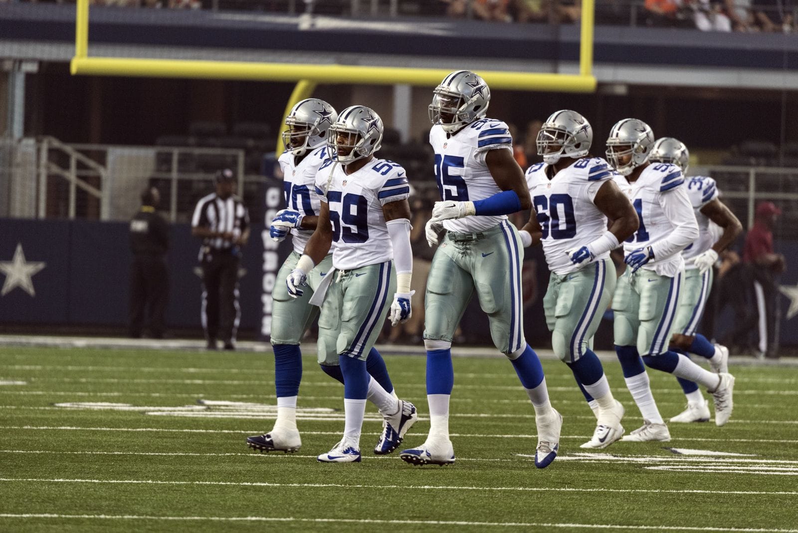 Dallas Cowboys defenders in white jerseys, silver-blue pants and silver helmets walking on the field at AT&T Stadium during a home game against the Denver Broncos in 2014