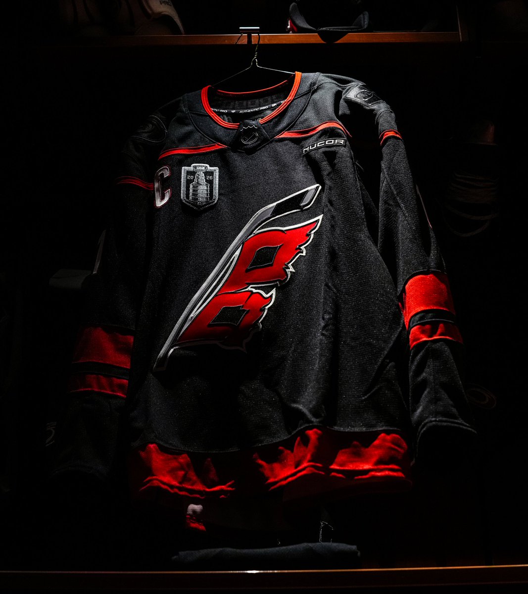 Close-up of a Carolina Hurricanes 2026 Stanley Cup Final jersey hanging in the Lenovo Center locker room with the new Stanley Cup Final 2026 patch on the right chest showing the silver Stanley Cup trophy silhouette with 20 on the left and 26 on the right flanking the Cup STANLEY CUP wordmark above and FINAL below, the captain C patch on the left chest, the Hurricanes red flag logo centered on the chest, and the NUCOR sponsor patch placement