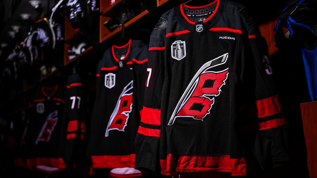 Carolina Hurricanes 2026 Stanley Cup Final jerseys hanging in the Lenovo Center locker room for Game 1 against the Vegas Golden Knights with the new Stanley Cup Final 2026 patch on the right chest, NUCOR sponsor patch, the Hurricanes red flag logo centered on the chest, the black home alternate sweater with red trim and red shoulder yoke
