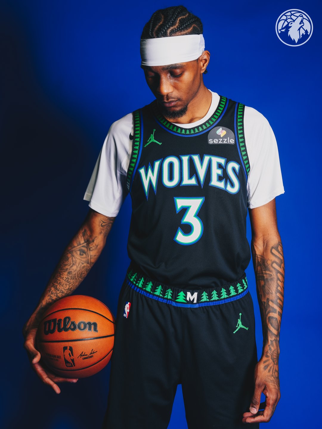 Minnesota Timberwolves new Statement Edition black alternate jersey 2026-27 standing pose on Jaden McDaniels number 3 with the green tree print pattern running across the collar trim and the shorts waistband, the WOLVES wordmark in bright blue with green outline, and the Jordan Brand jumpman patch on the chest