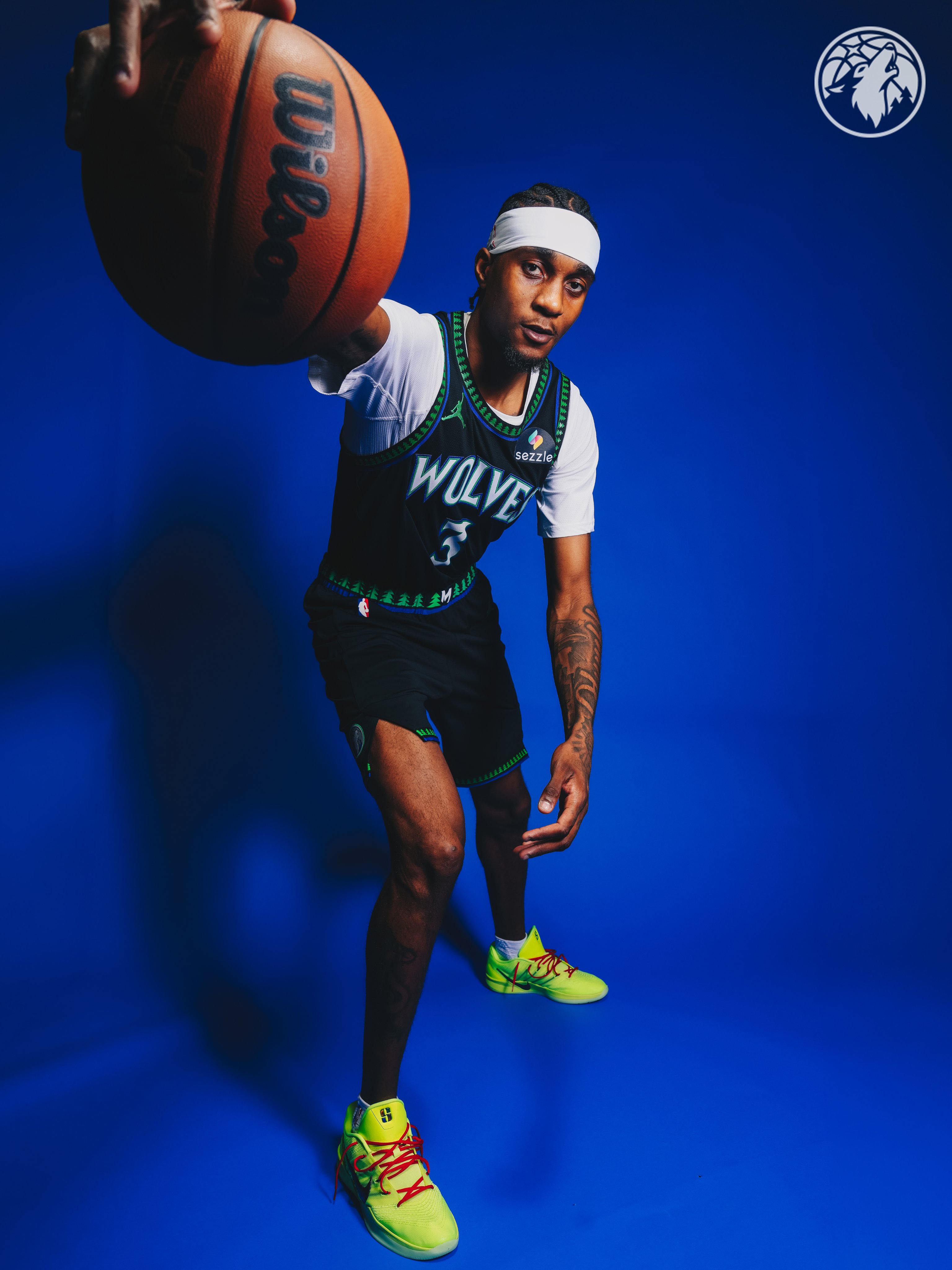 Minnesota Timberwolves new Statement Edition black alternate jersey 2026-27 dynamic pose on Jaden McDaniels showing the full uniform in motion with the throwback green tree pattern collar trim, the WOLVES blue wordmark, the matching black shorts with green Jumpman, and the green tree print also wrapping the shorts waistband