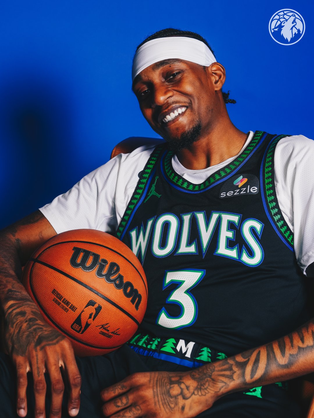 Minnesota Timberwolves new Statement Edition black alternate jersey 2026-27 close-up seated pose on Jaden McDaniels showing the green tree pattern wrapping the collar and armhole trim, the WOLVES wordmark in bright blue with the green outline, the Sezzle jersey patch on the right chest, and the Jordan jumpman on the left chest