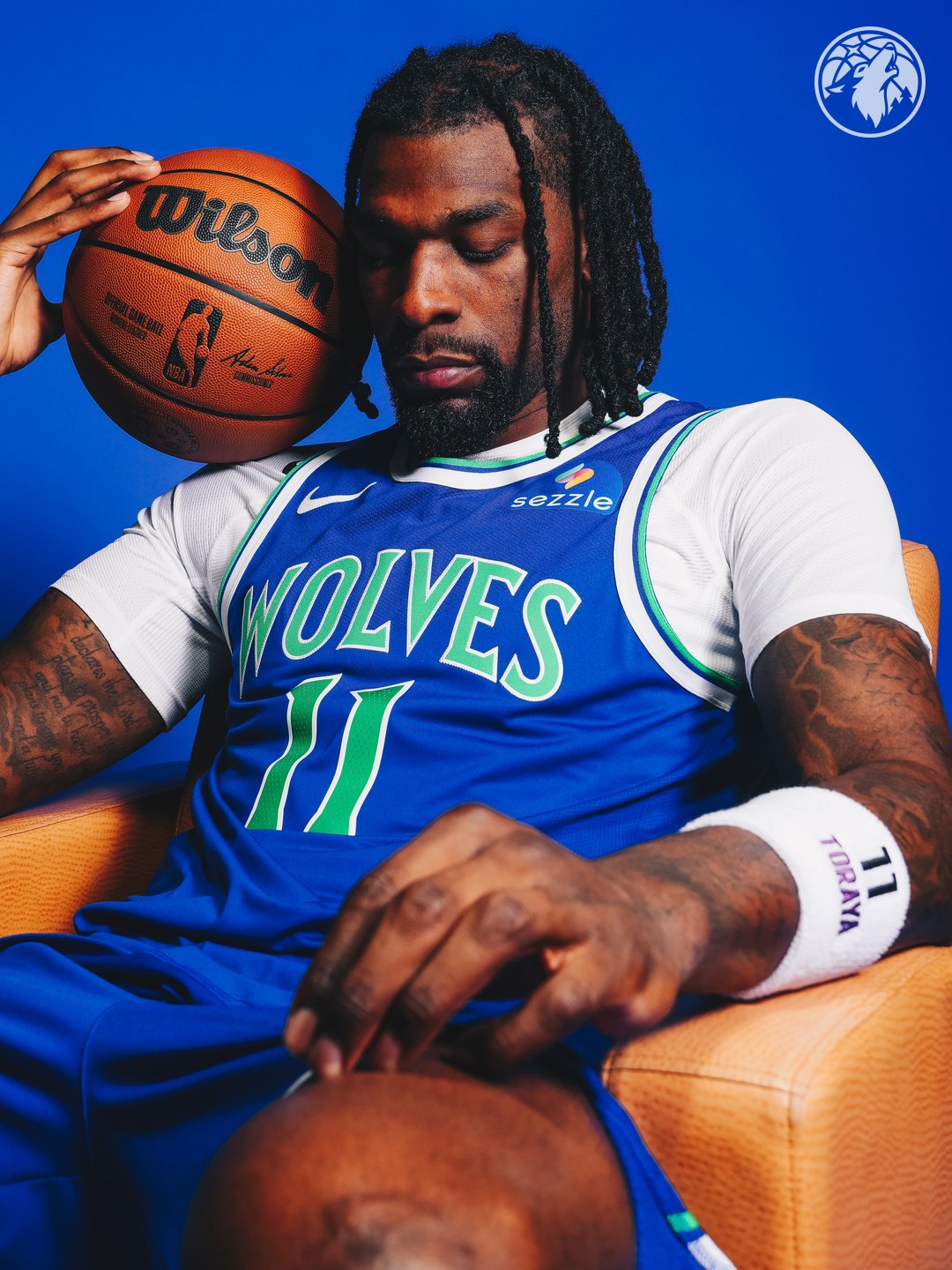 Minnesota Timberwolves new Icon Edition blue road jersey 2026-27 close-up seated pose on Naz Reid with the basketball at his head, showing the WOLVES throwback wordmark in bright green, the #11 chest number, the Sezzle jersey patch on the right chest, and the embroidered green outline on the throwback typography