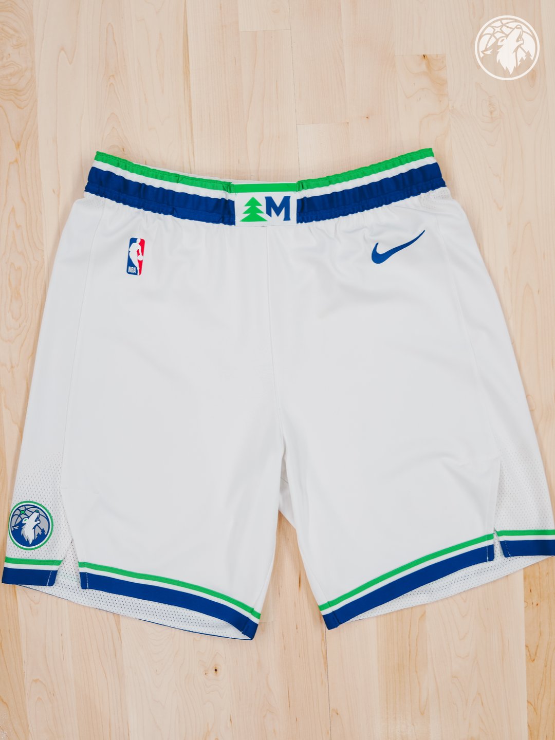 Minnesota Timberwolves new Association Edition white home shorts 2026-27 laid flat product shot showing the green tree and Minnesota M monogram waistband label, the green and blue piping at the hem, and the new wolf head crest patch on the side leg