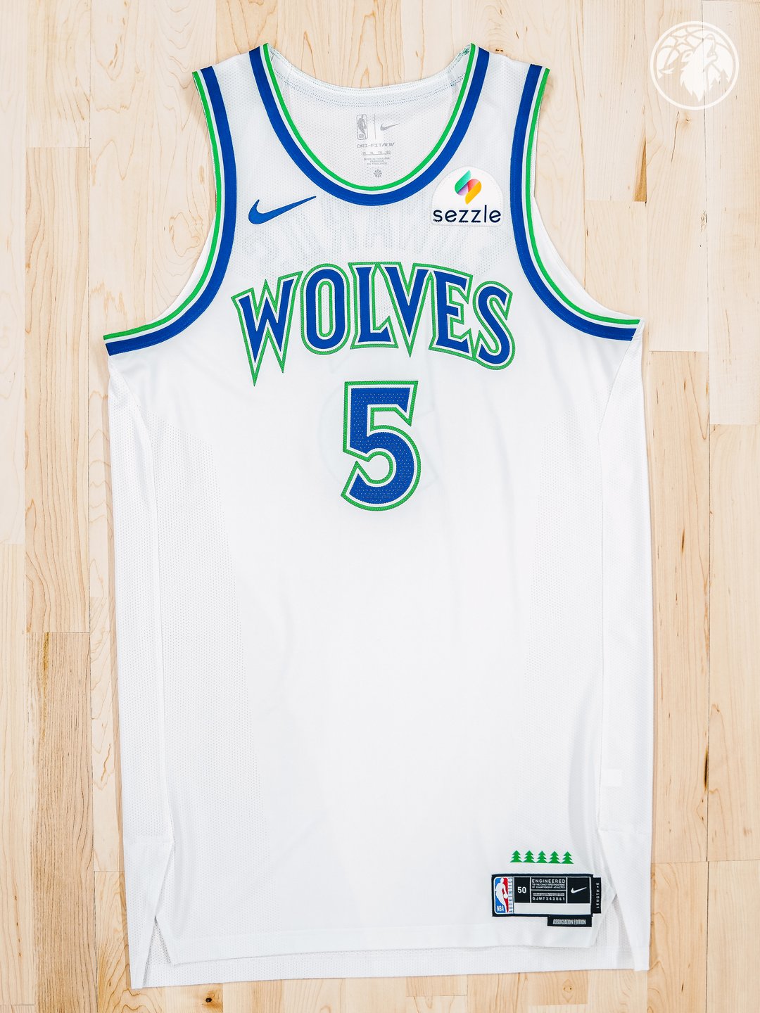 Minnesota Timberwolves new Association Edition white home jersey 2026-27 laid flat product shot on a wood floor showing the full front view with the WOLVES throwback wordmark in blue and green outline, the #5 chest number for Anthony Edwards, the green tree silhouettes on the lower tag, and the Sezzle jersey patch on the right chest