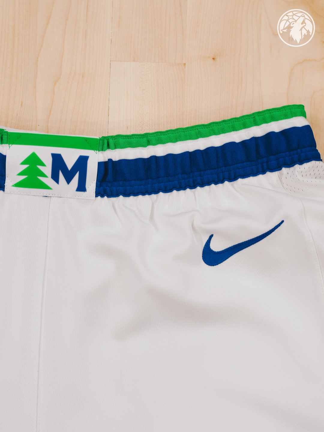 Minnesota Timberwolves new Association Edition white home shorts 2026-27 waistband macro close-up showing the green pine tree silhouette next to the blue Minnesota M monogram label, the bright green stripe trim above the blue waistband, and the embroidered Nike swoosh on the leg