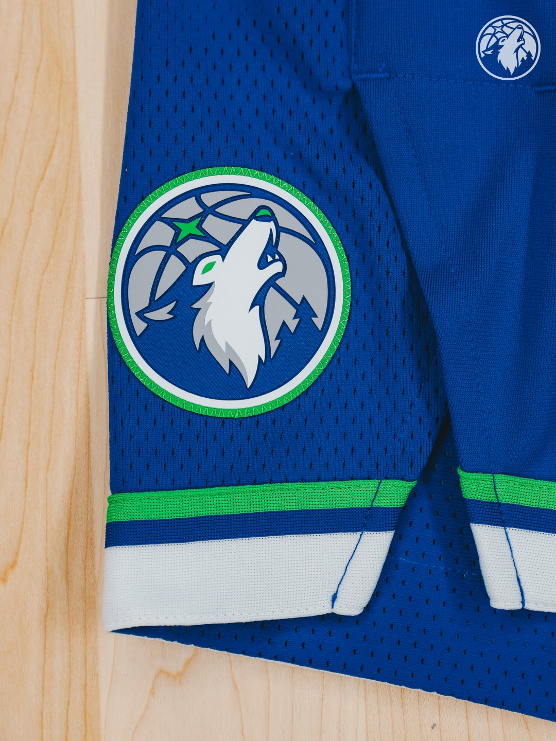 Minnesota Timberwolves new Icon Edition blue road jersey 2026-27 side detail close-up showing the new wolf head crest patch on the side panel, the green and white striped hem trim at the bottom of the jersey, and the saturated brighter blue jersey body