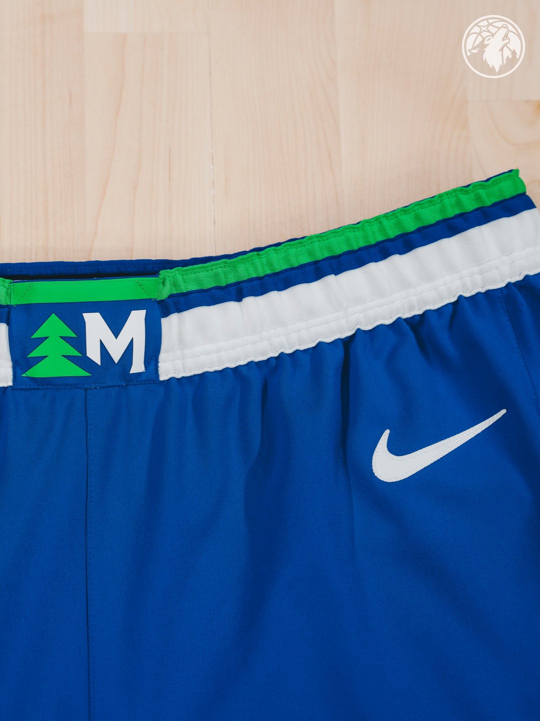 Minnesota Timberwolves new Icon Edition blue road shorts 2026-27 waistband macro close-up showing the green pine tree silhouette next to the white Minnesota M monogram label, the bright green stripe trim above the white waistband stripe, and the embroidered white Nike swoosh on the saturated blue shorts