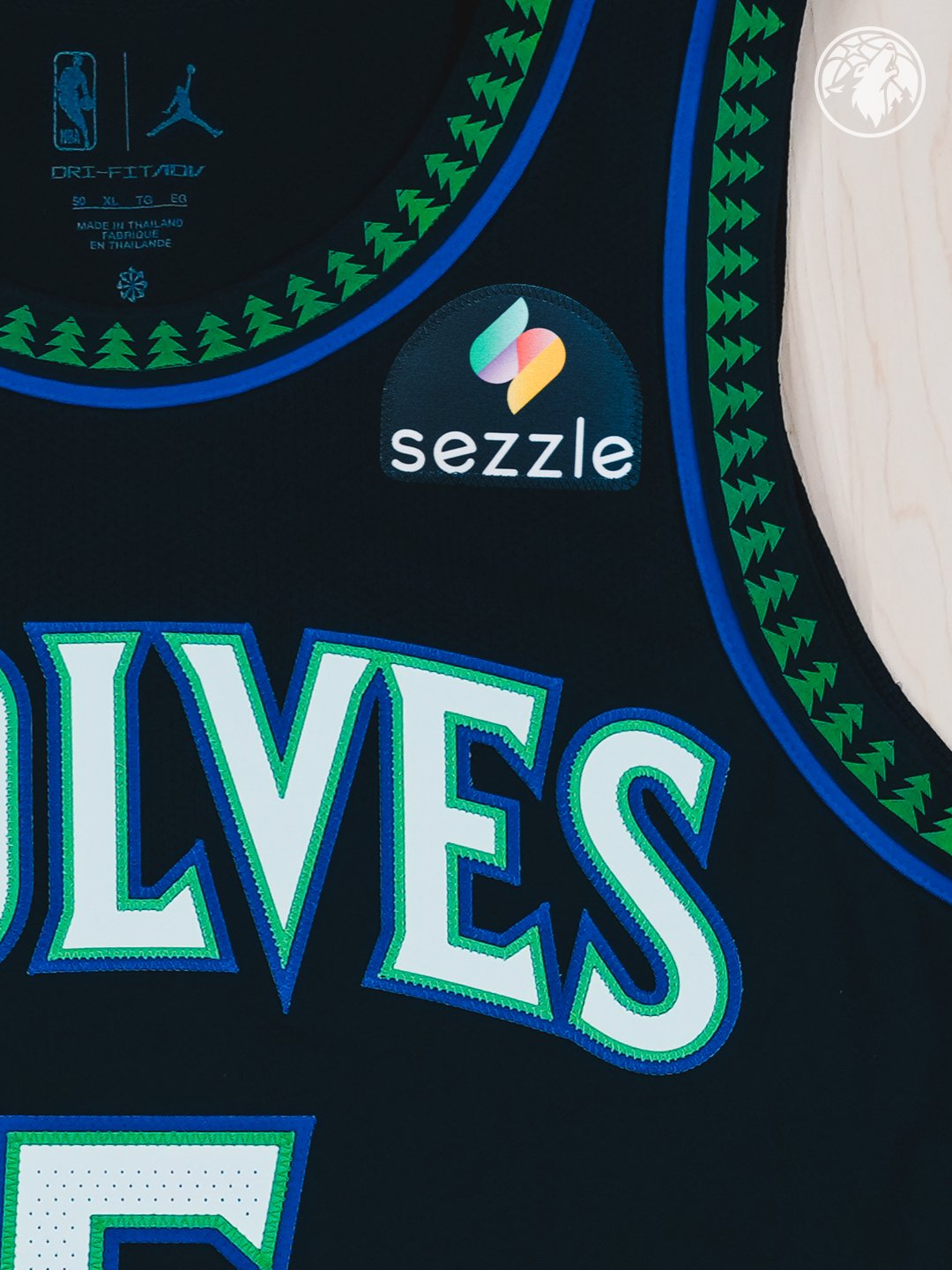 Minnesota Timberwolves new Statement Edition black alternate jersey 2026-27 macro close-up of the WOLVES wordmark and Sezzle jersey patch showing the green tree pattern wrapping the collar and shoulder yoke with the blue outline detail, the embroidered blue and green letterforms, and the Hardwood Classic side panel motif returning as the centerpiece of the Wolves Statement Edition