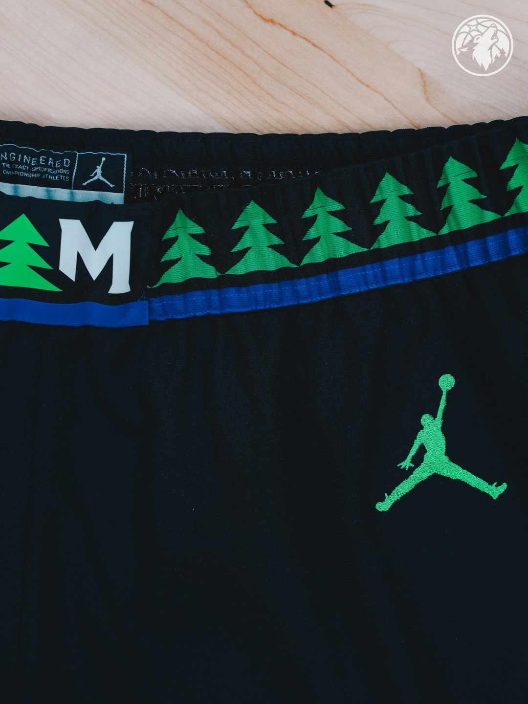 Minnesota Timberwolves new Statement Edition black alternate shorts 2026-27 waistband macro close-up showing the row of green pine tree silhouettes running across the waistband, the white Minnesota M monogram label, the bright green Jordan Brand Jumpman on the leg, and the bright blue stripe trim at the waistband edge