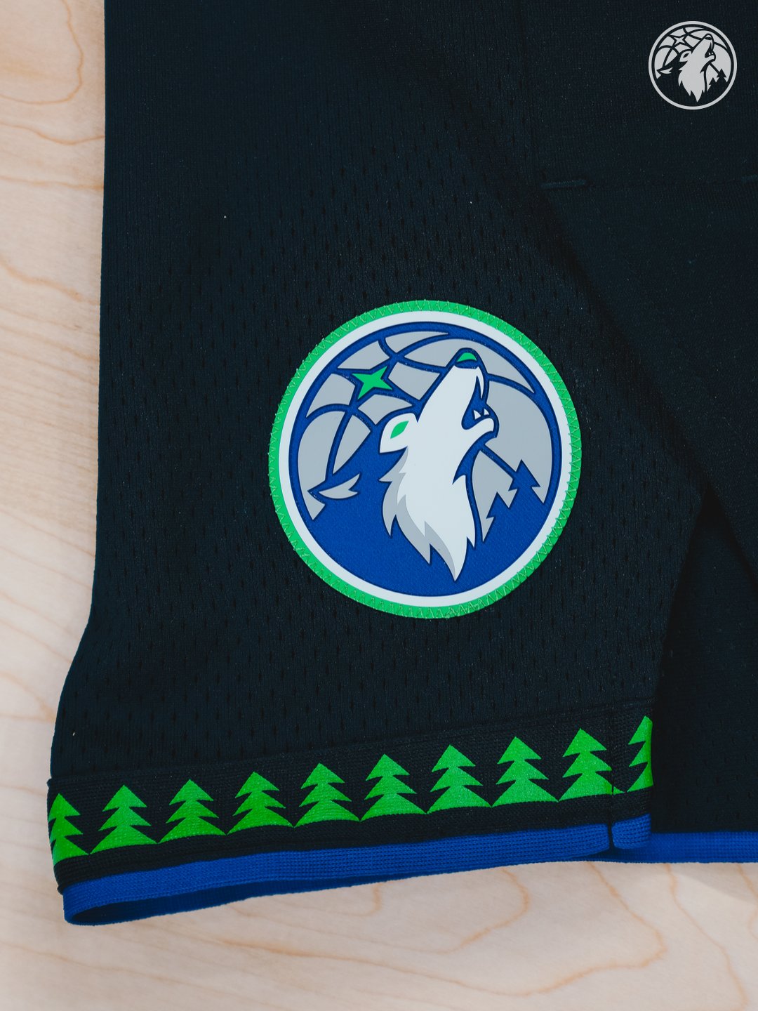 Minnesota Timberwolves new Statement Edition black alternate shorts 2026-27 side leg detail showing the new wolf head crest patch on the side panel, the green tree silhouette pattern running across the bottom hem of the shorts, and the bright blue stripe trim at the very edge of the shorts
