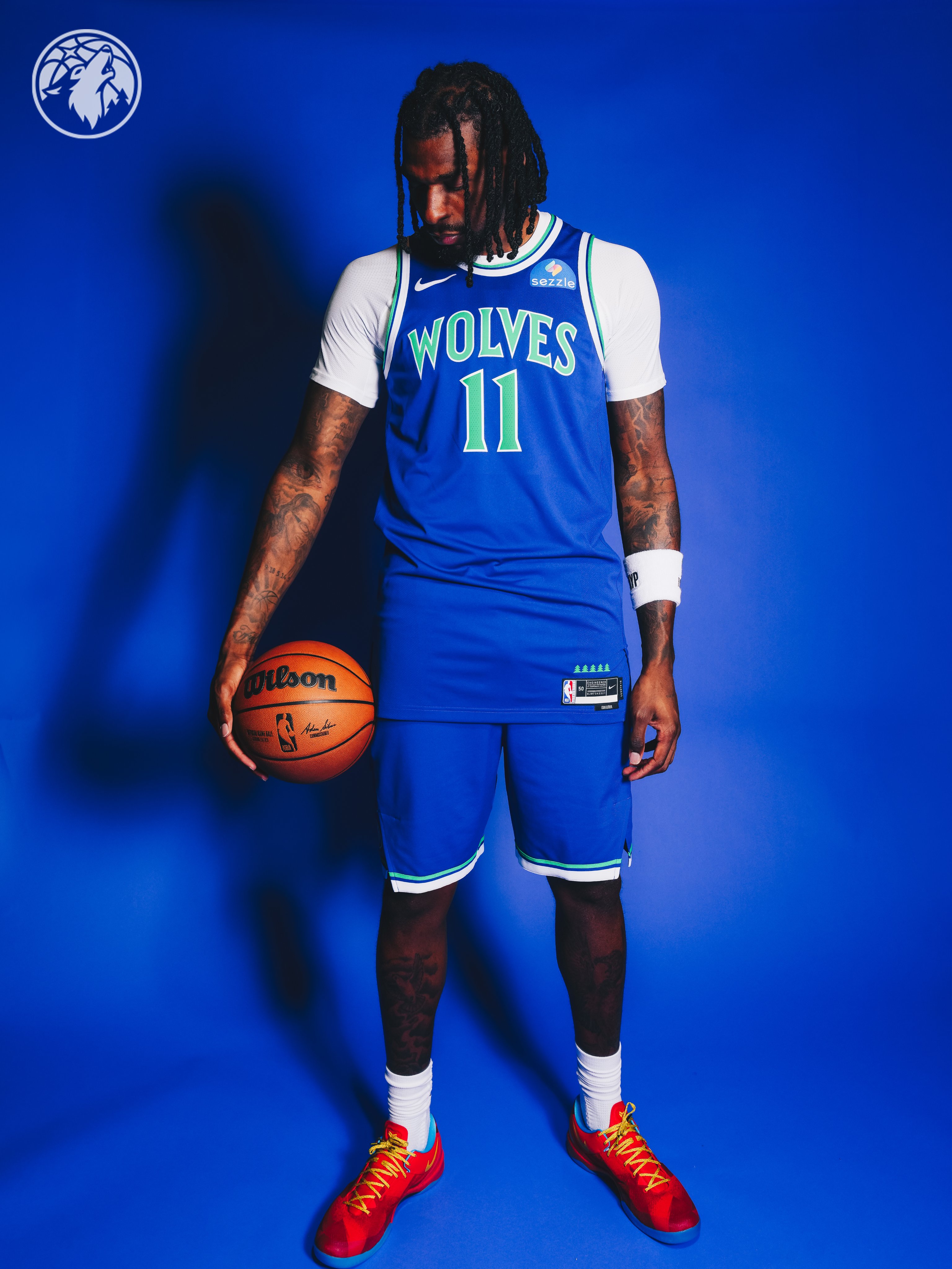 Minnesota Timberwolves new 2026-27 Icon Edition blue road jersey officially released and modeled by Naz Reid number 11 standing in front of a saturated blue backdrop with the new wolf head crest logo in the top left corner, the brighter blue and green throwback color palette returning from the original 1989 expansion identity, the WOLVES wordmark in bright green across the chest, and the green and white piping on the matching blue road shorts